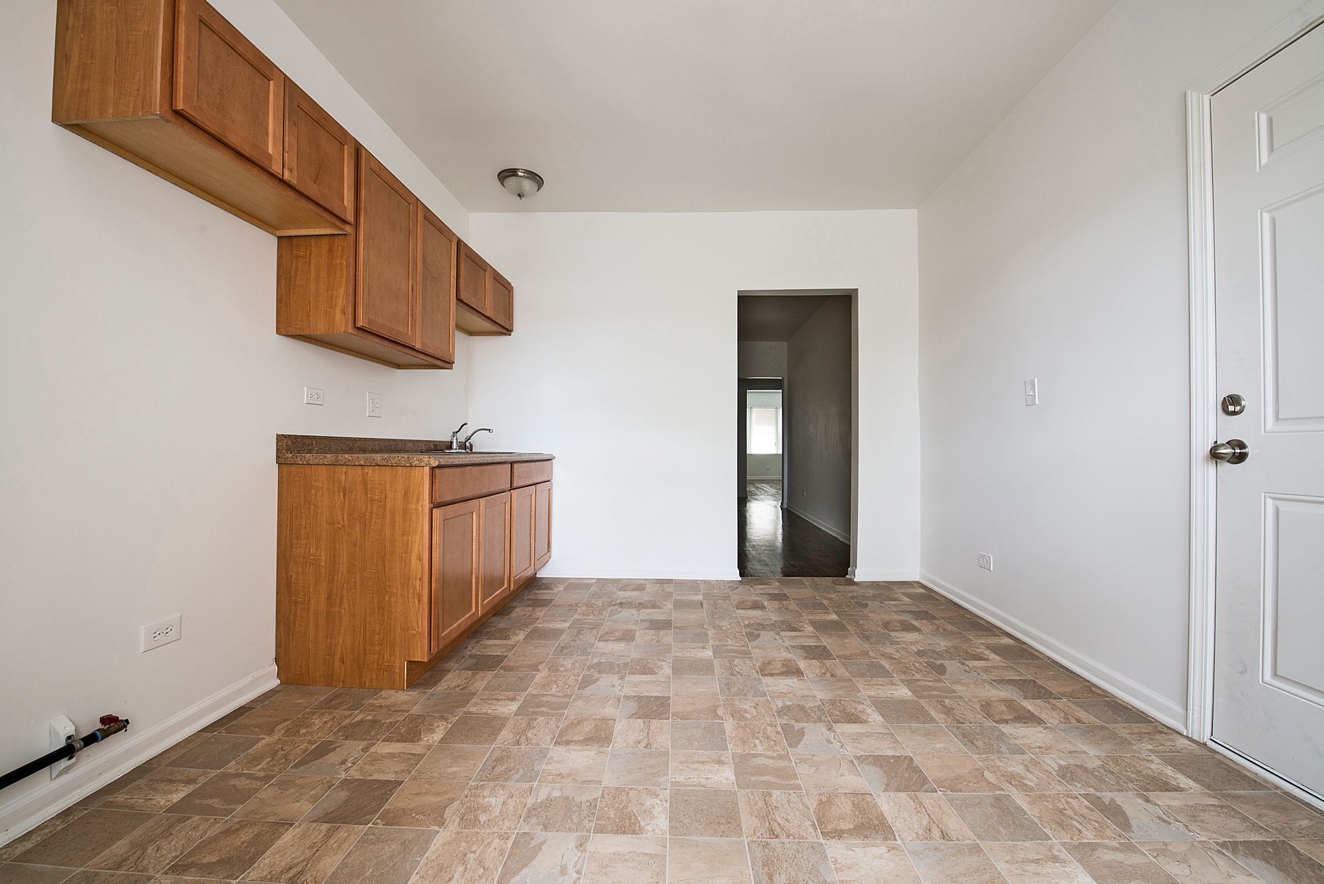Empty kitchen with brown cabinets, countertop, and patterned floor. White walls, door, and a hallway entrance.