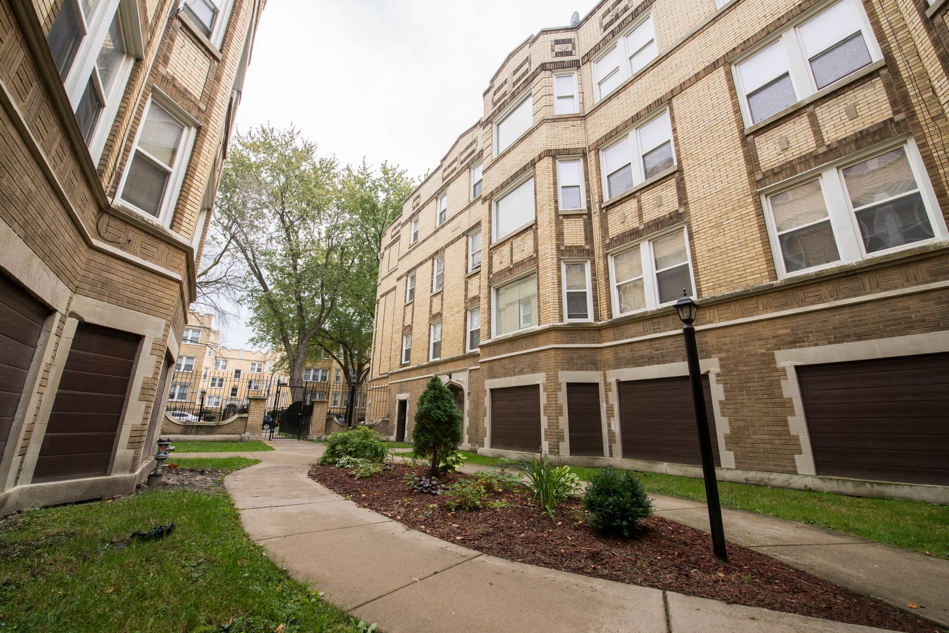 Apartment complex exterior, brick buildings, garages, walkway, grass, cloudy sky.