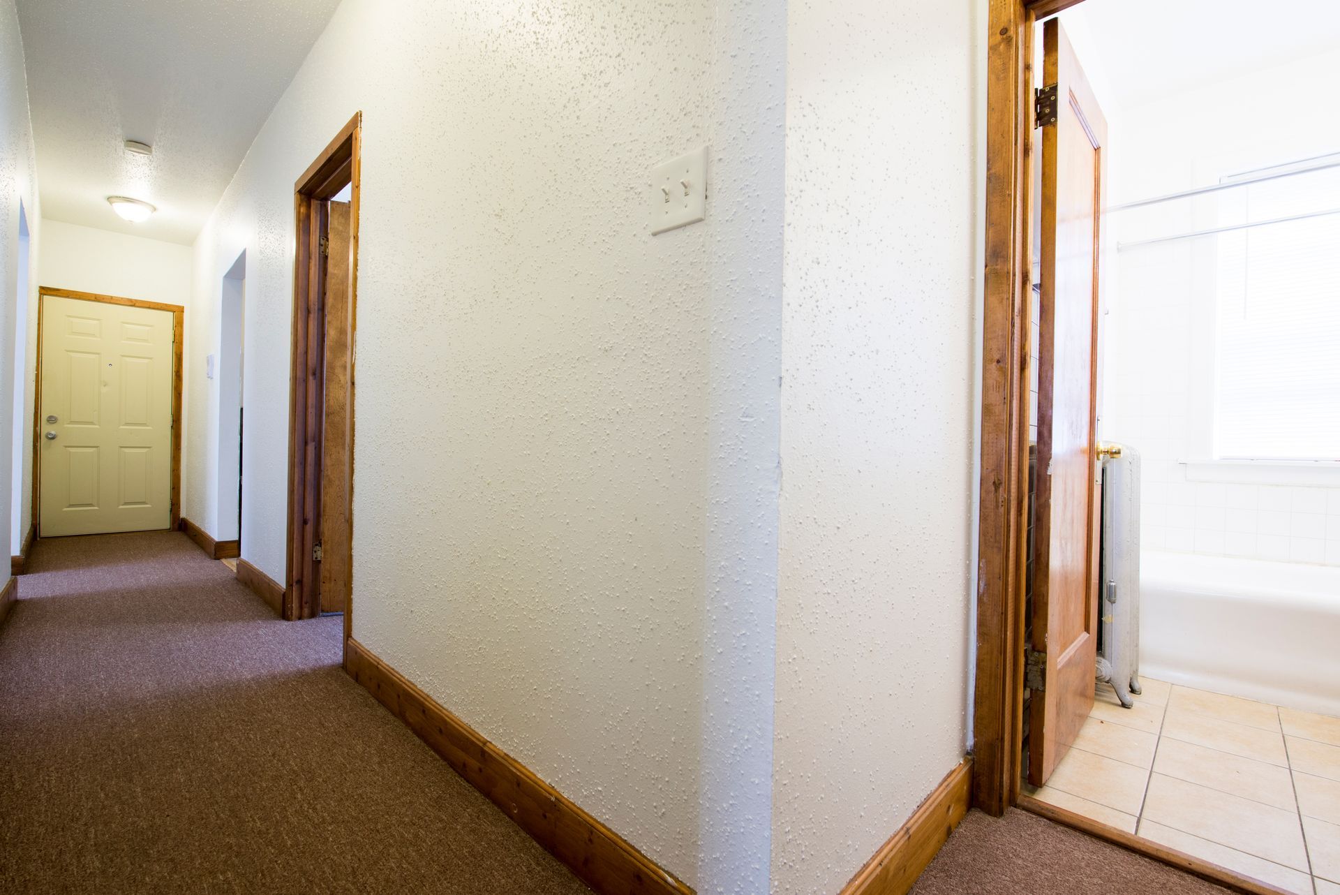 Hallway with doors, beige carpet, and textured white walls. One door is open to a bathroom.