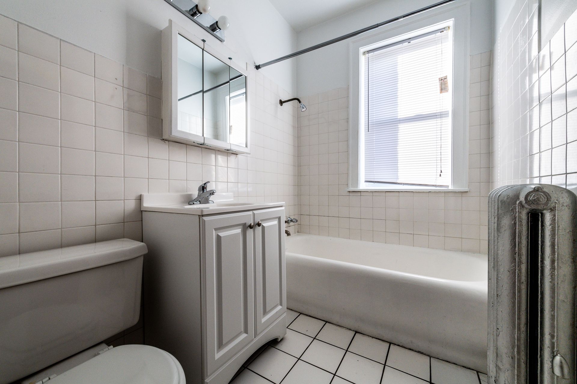Bathroom with white tile walls, toilet, vanity, bathtub, and window with a curtain.