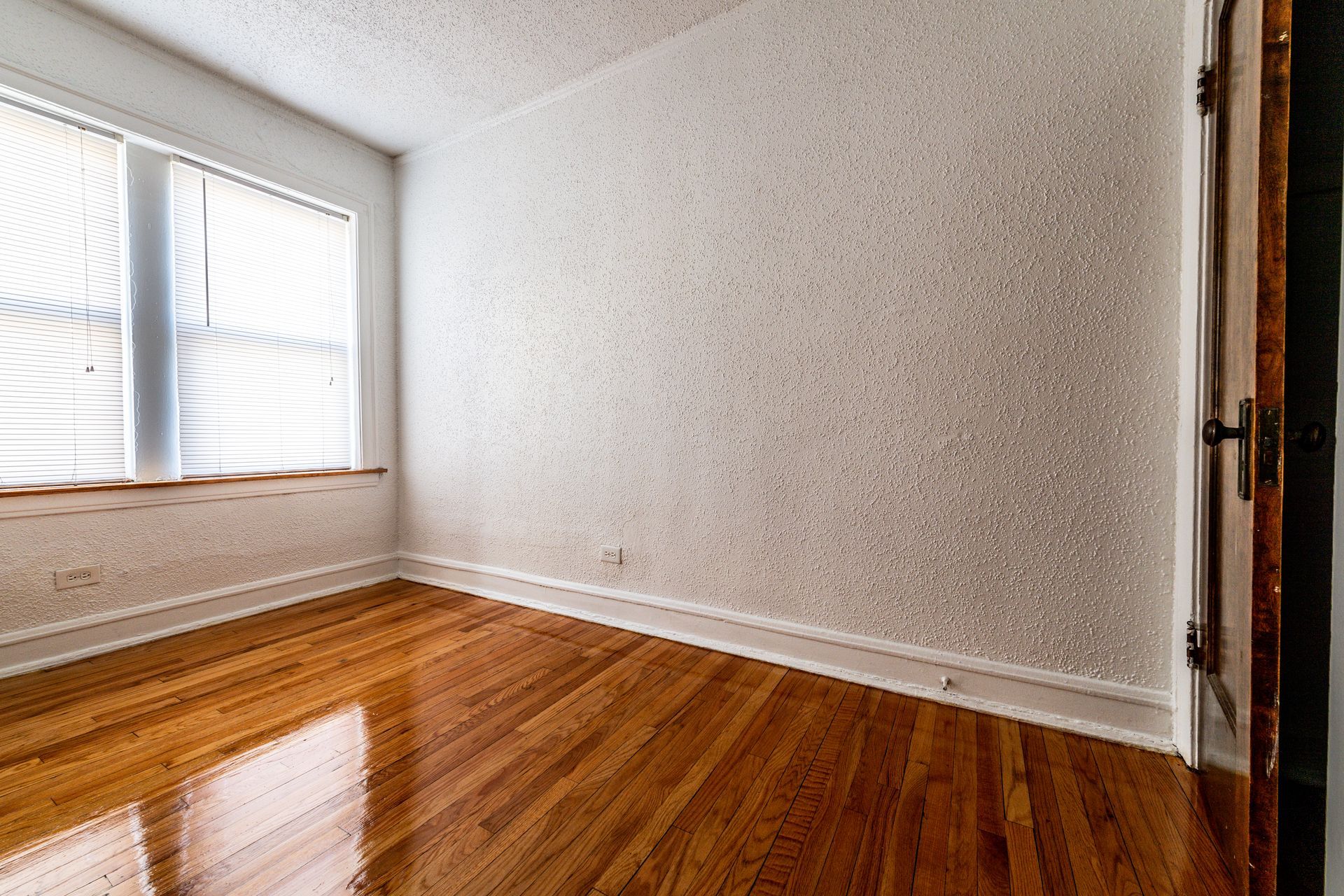 Empty room with wood floors, textured white walls, a window with blinds, and a doorway.