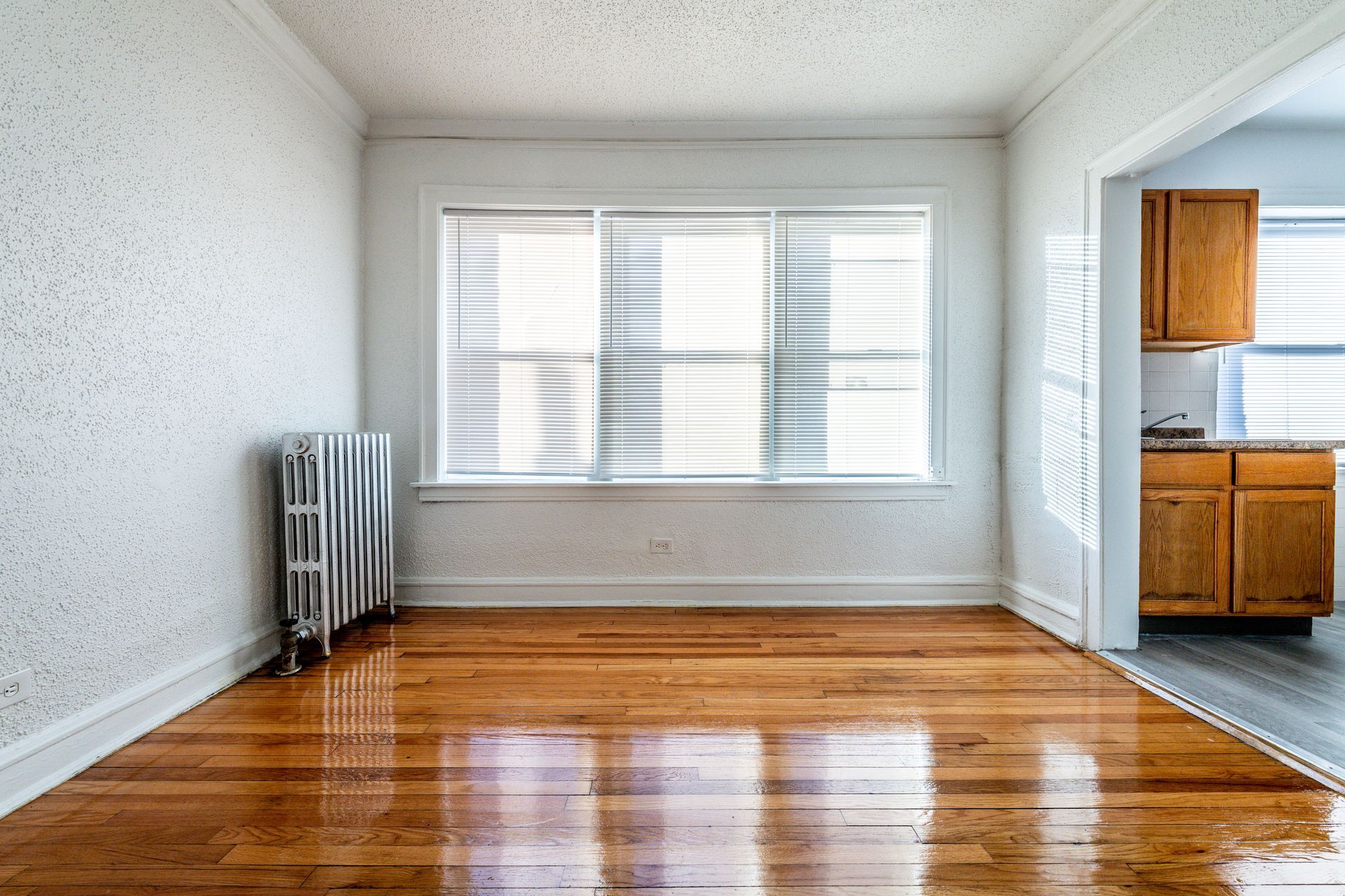 Empty room with hardwood floors, a radiator, and a window with blinds; kitchen visible in the doorway.