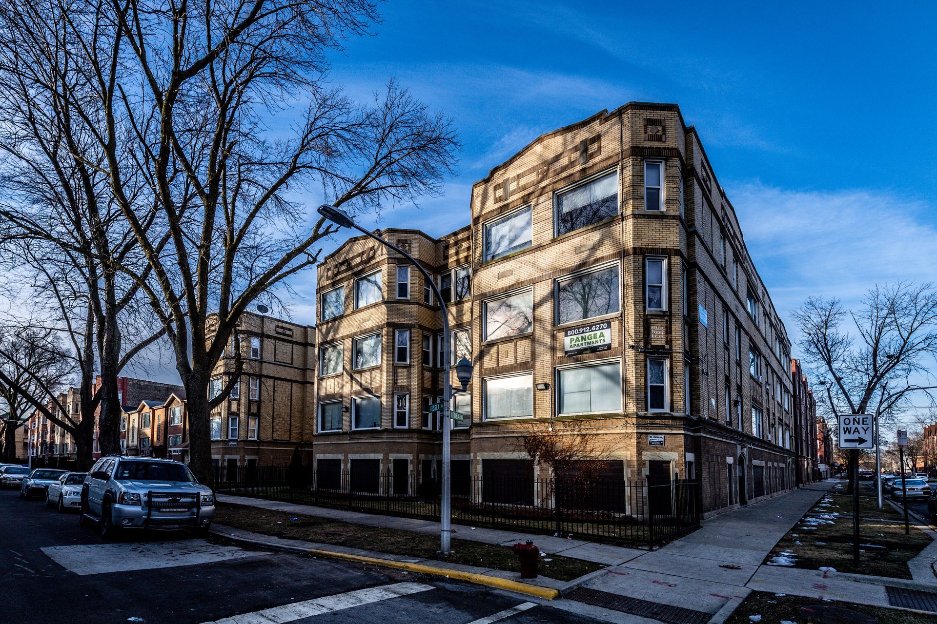 Three-story brick apartment building on a city street; bare trees, blue sky, cars parked along the street.