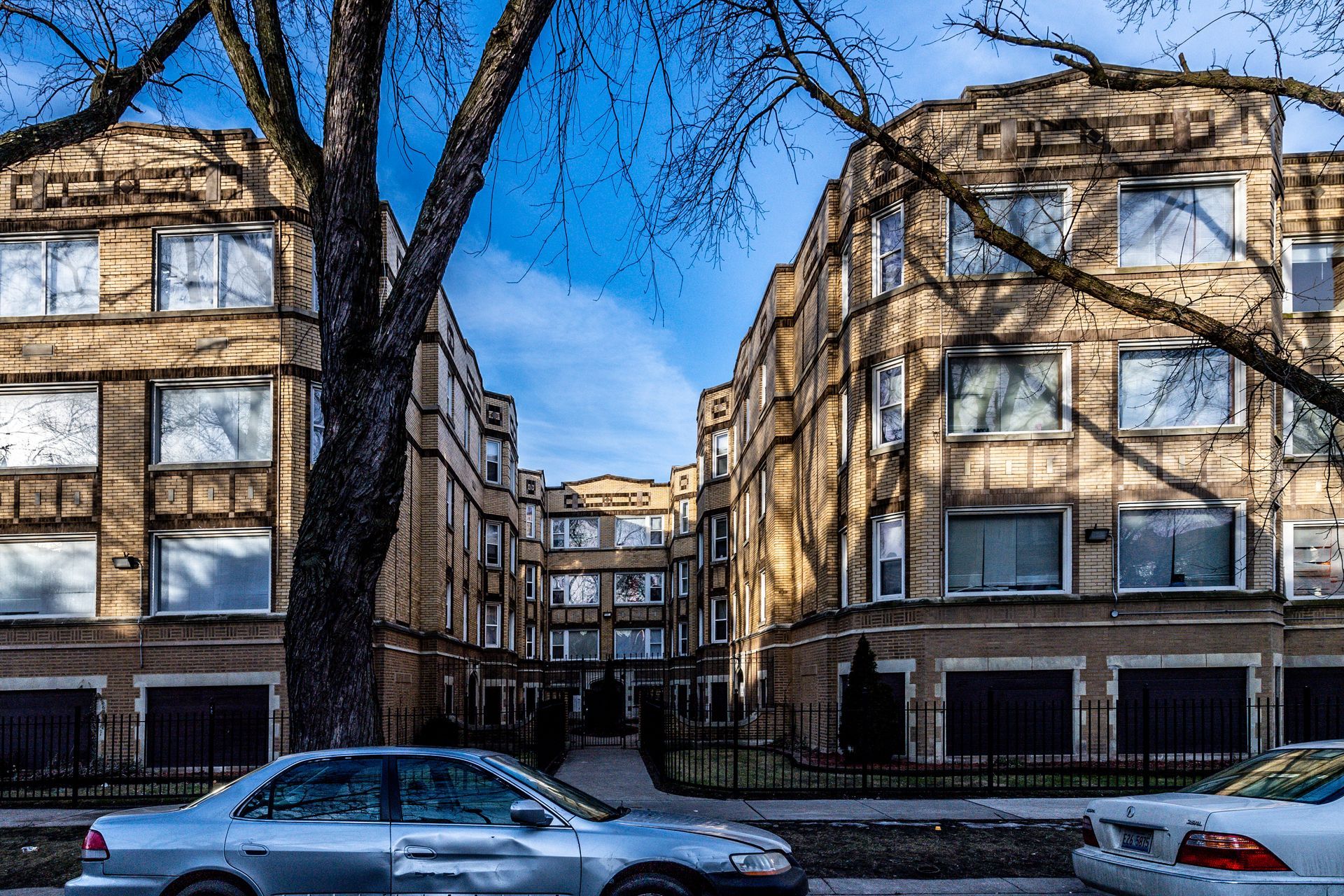Apartment buildings with beige brick facades and windows, trees in the foreground, and a car parked on the street.