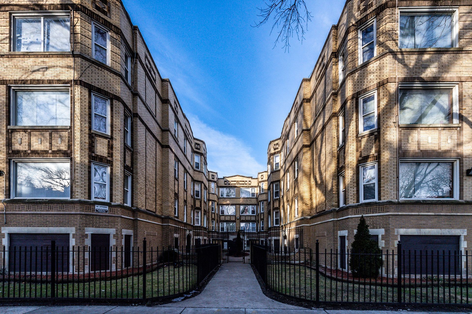 Two tan brick apartment buildings face each other, connected by a walkway and gate. Blue sky overhead.