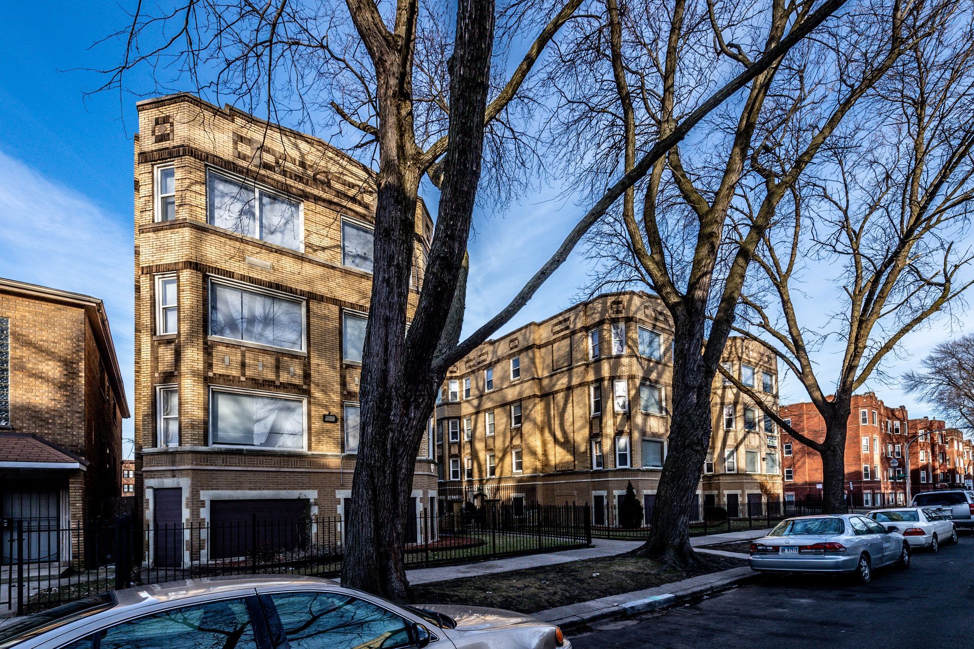 A three-story, beige apartment building on a tree-lined street with parked cars.