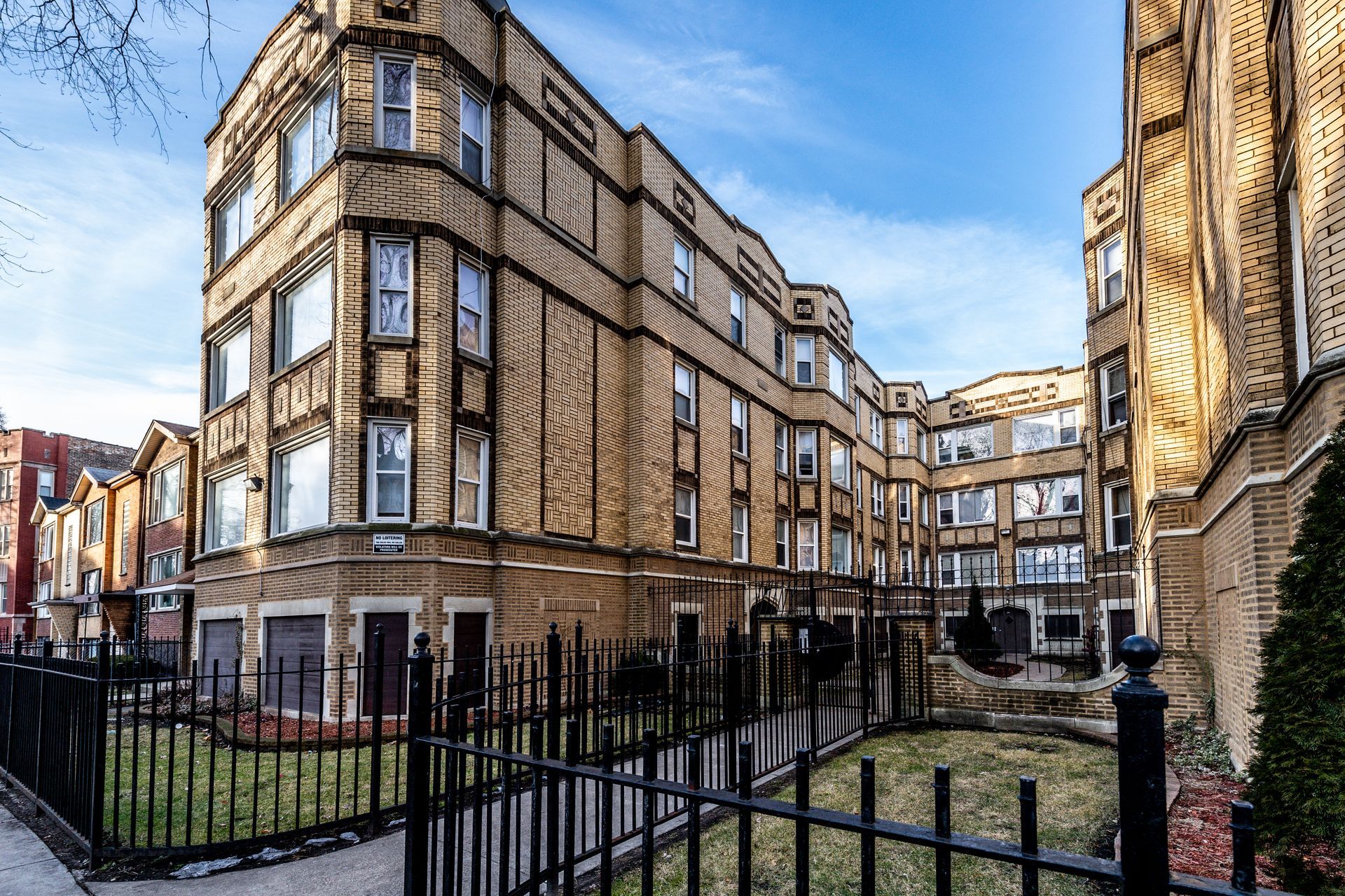 Multi-story brick apartment building with black fence and small yard.
