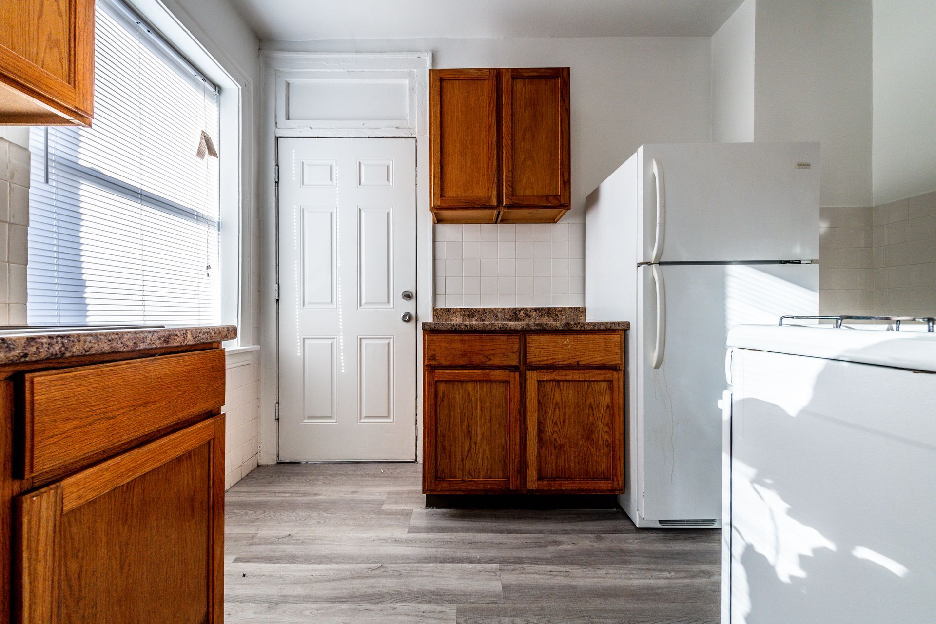 Kitchen with wooden cabinets, white appliances, and a door, natural light streaming in from the window.