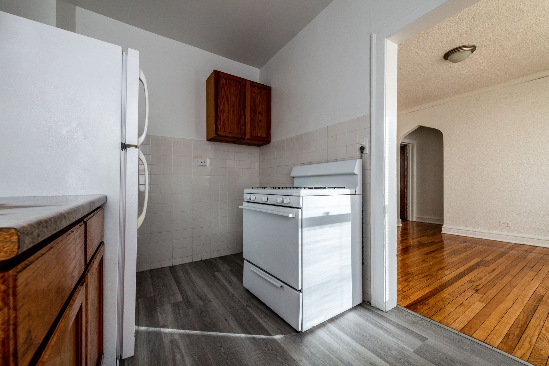 Kitchen with white appliances, wooden cabinets, and gray flooring; doorway to a hardwood floor room.