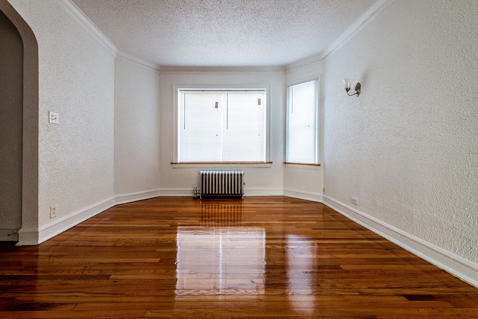 Empty room with hardwood floors, white textured walls, and windows with blinds.