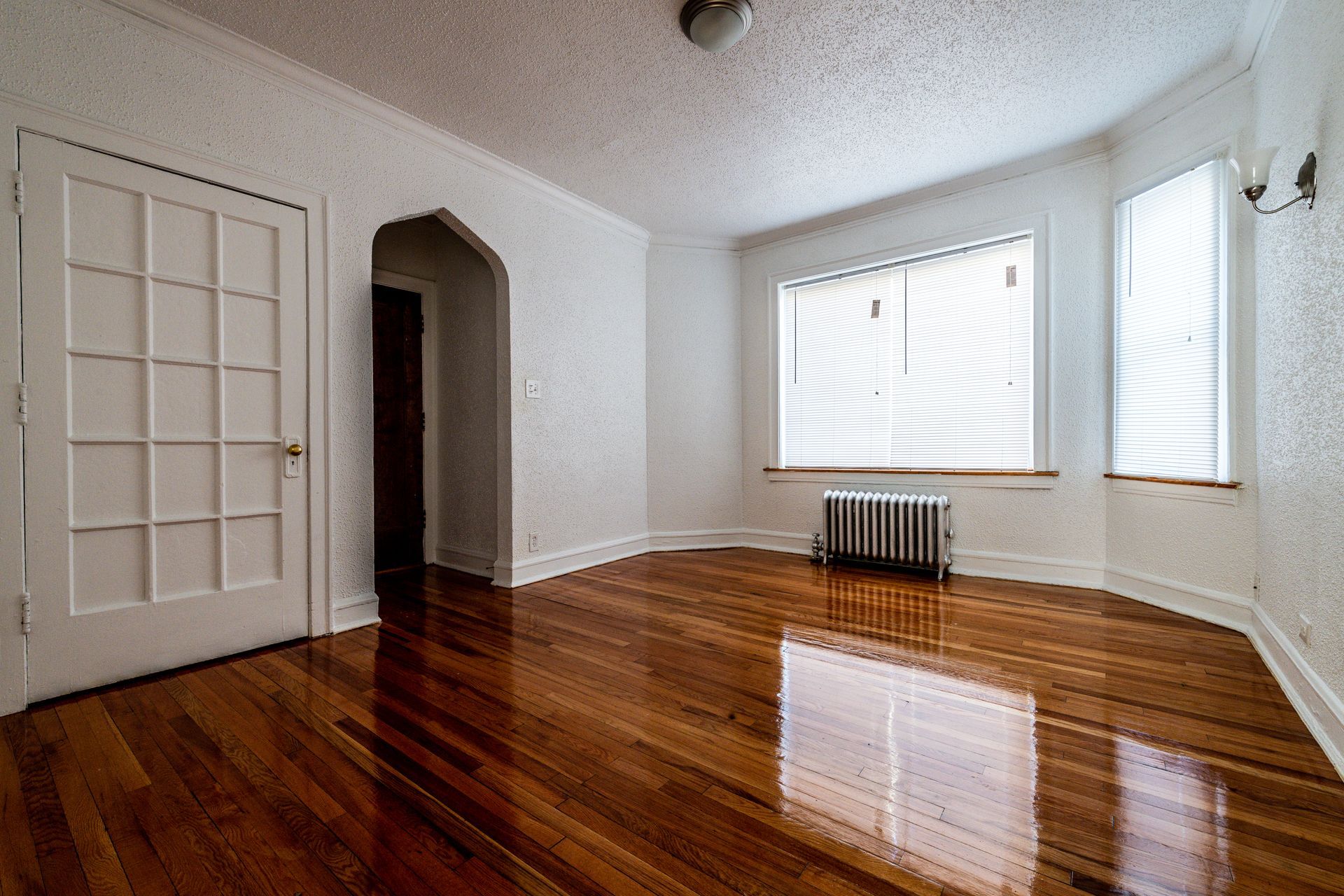 Empty room with wood floors, radiator, and arched doorway. Two windows with blinds. White walls and door.