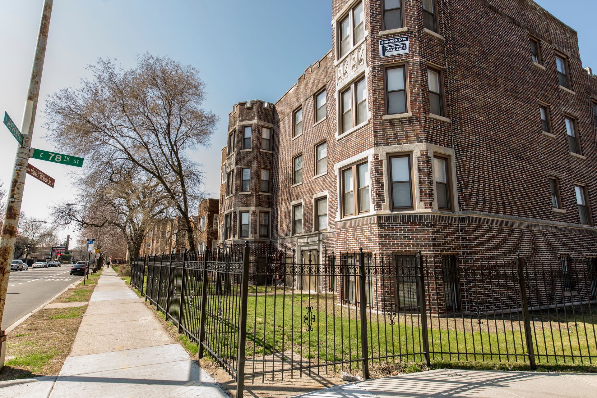 Brick apartment building with decorative turrets and wrought iron fence along a sidewalk.