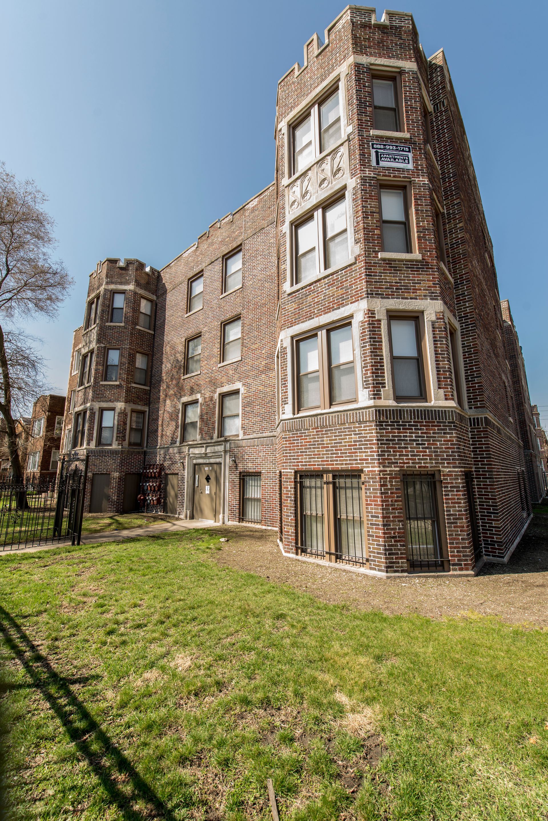 Brick apartment building with turret-like corners on a sunny day. Green lawn in front.