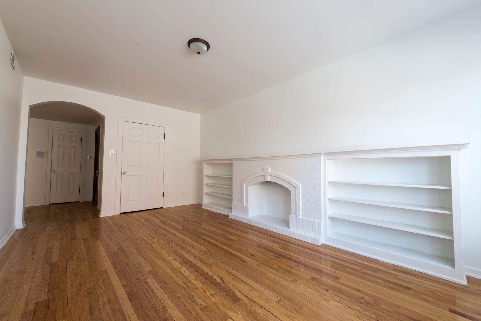 Empty room with hardwood floors, built-in shelves, and a faux fireplace. White walls and door.