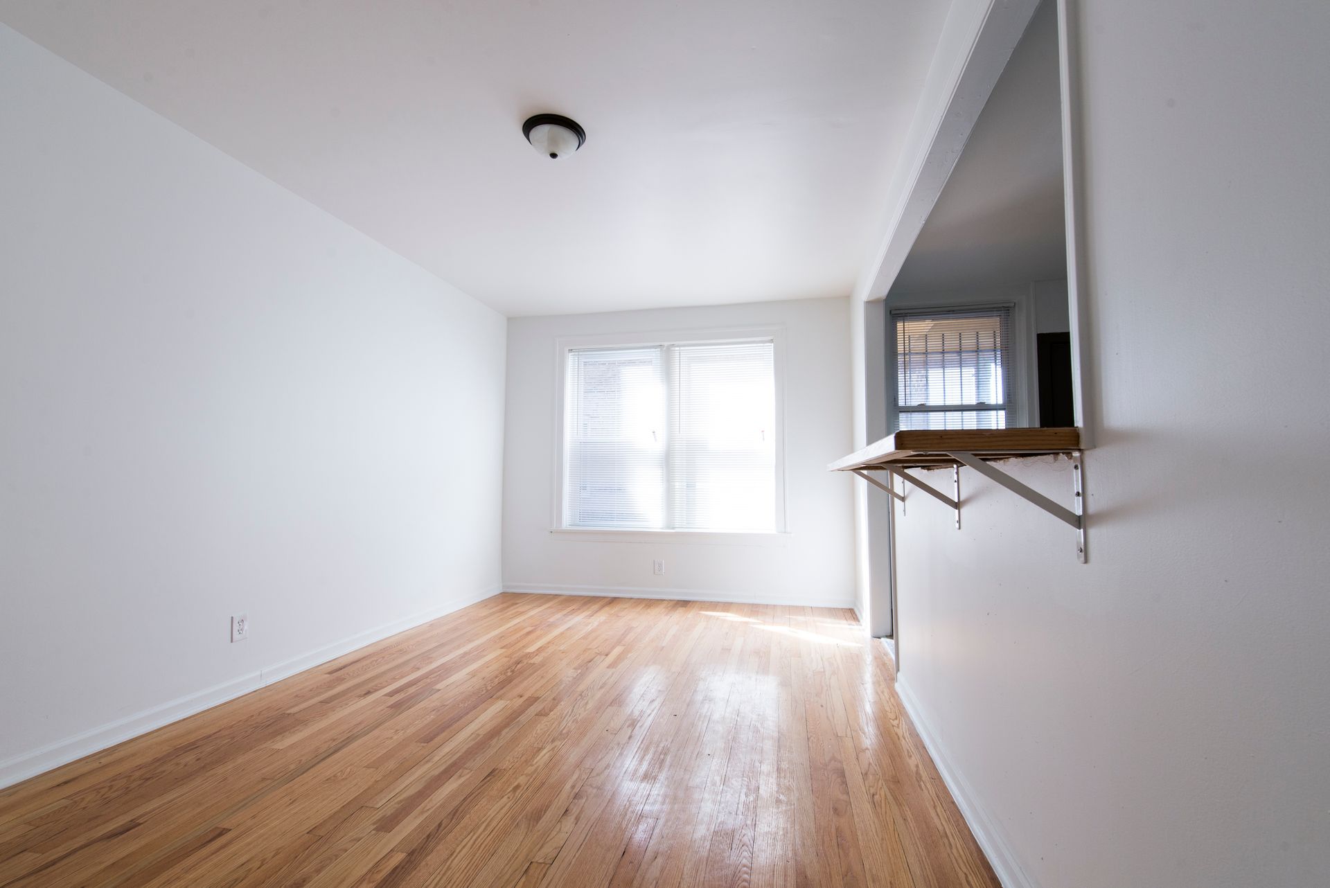 Empty room with hardwood floors, white walls, and a window with blinds. A pass-through to another room is on the right.
