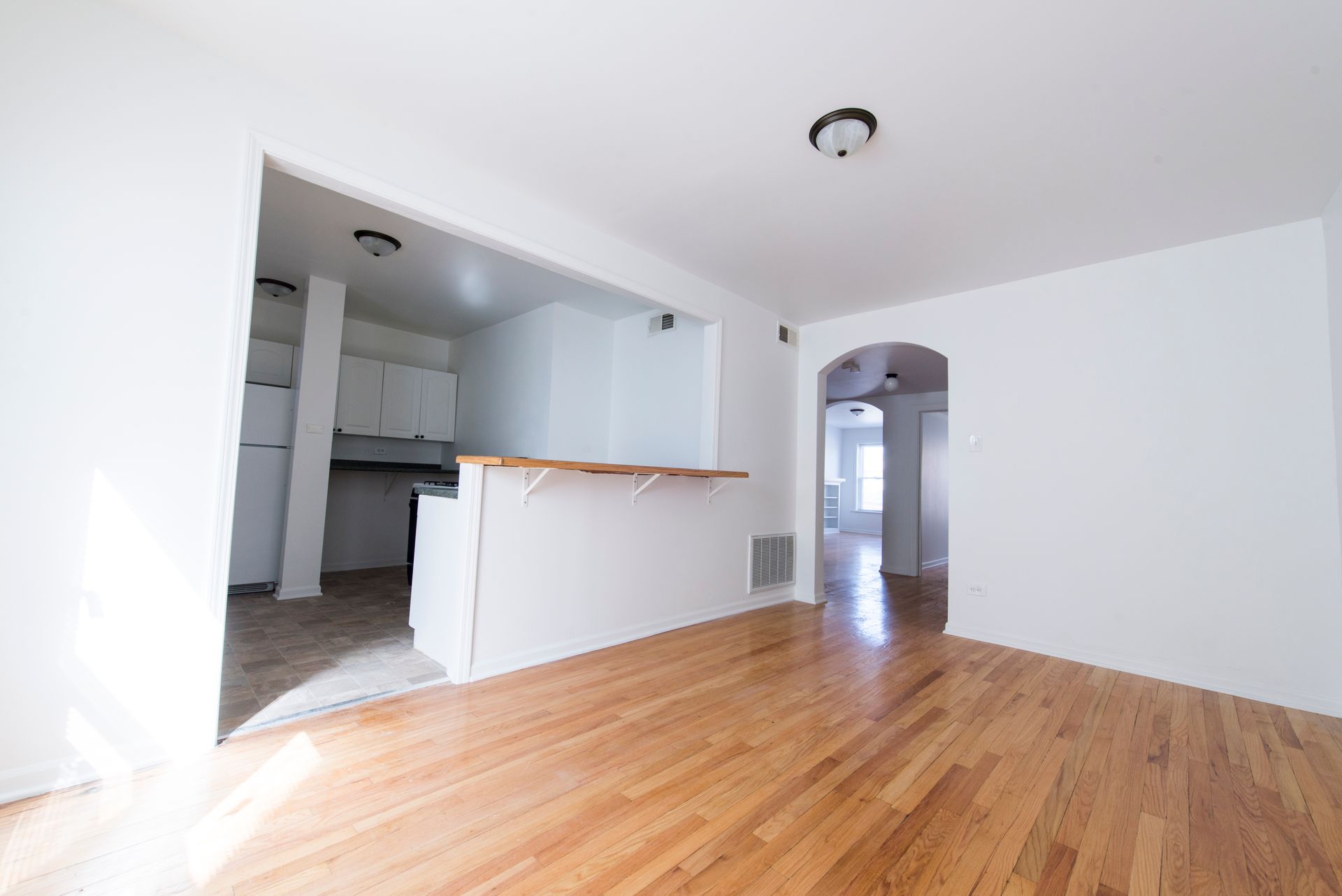 Empty living room with hardwood floors, a kitchen opening, and a doorway.