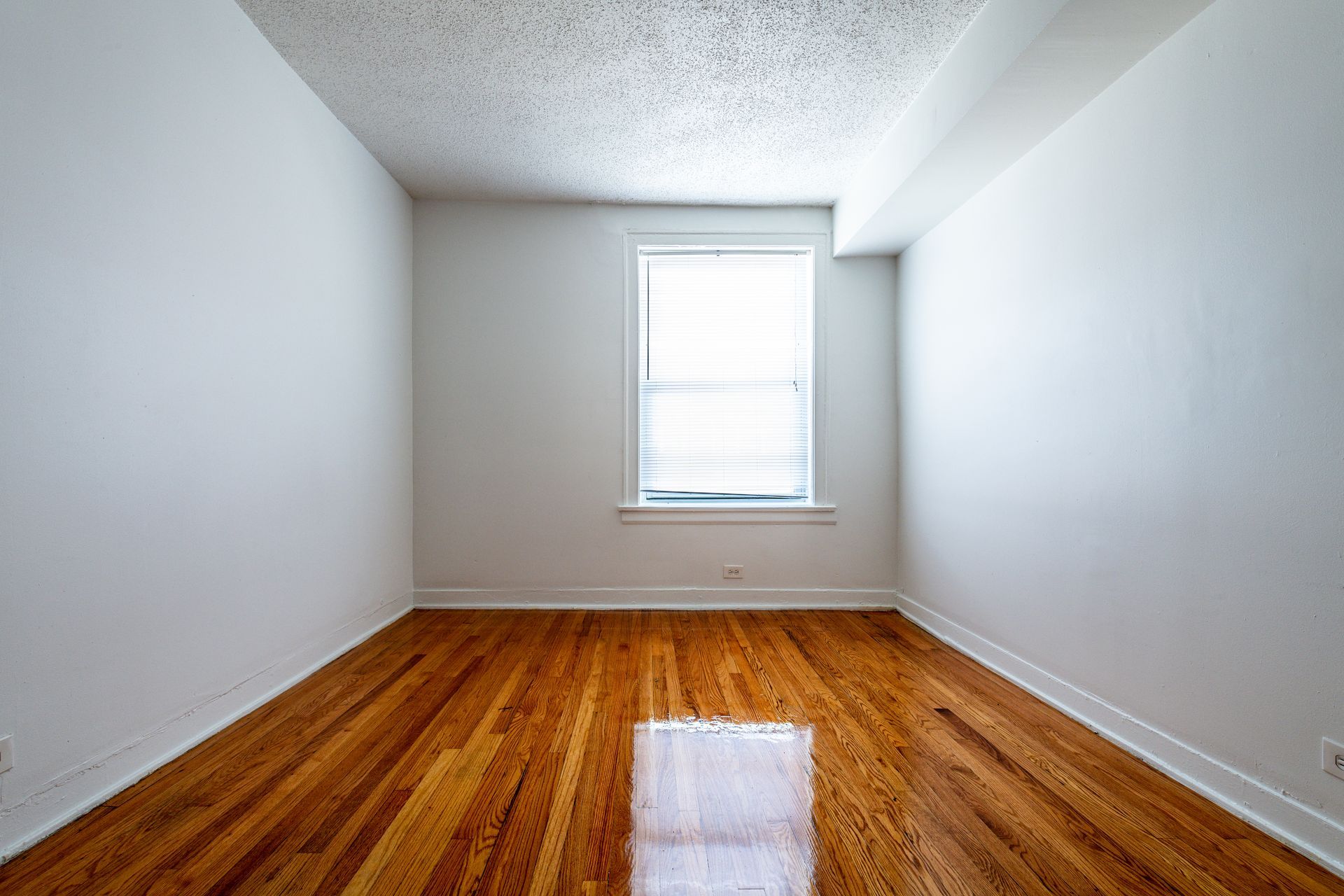Empty room with hardwood floors, white walls, and a window with blinds.
