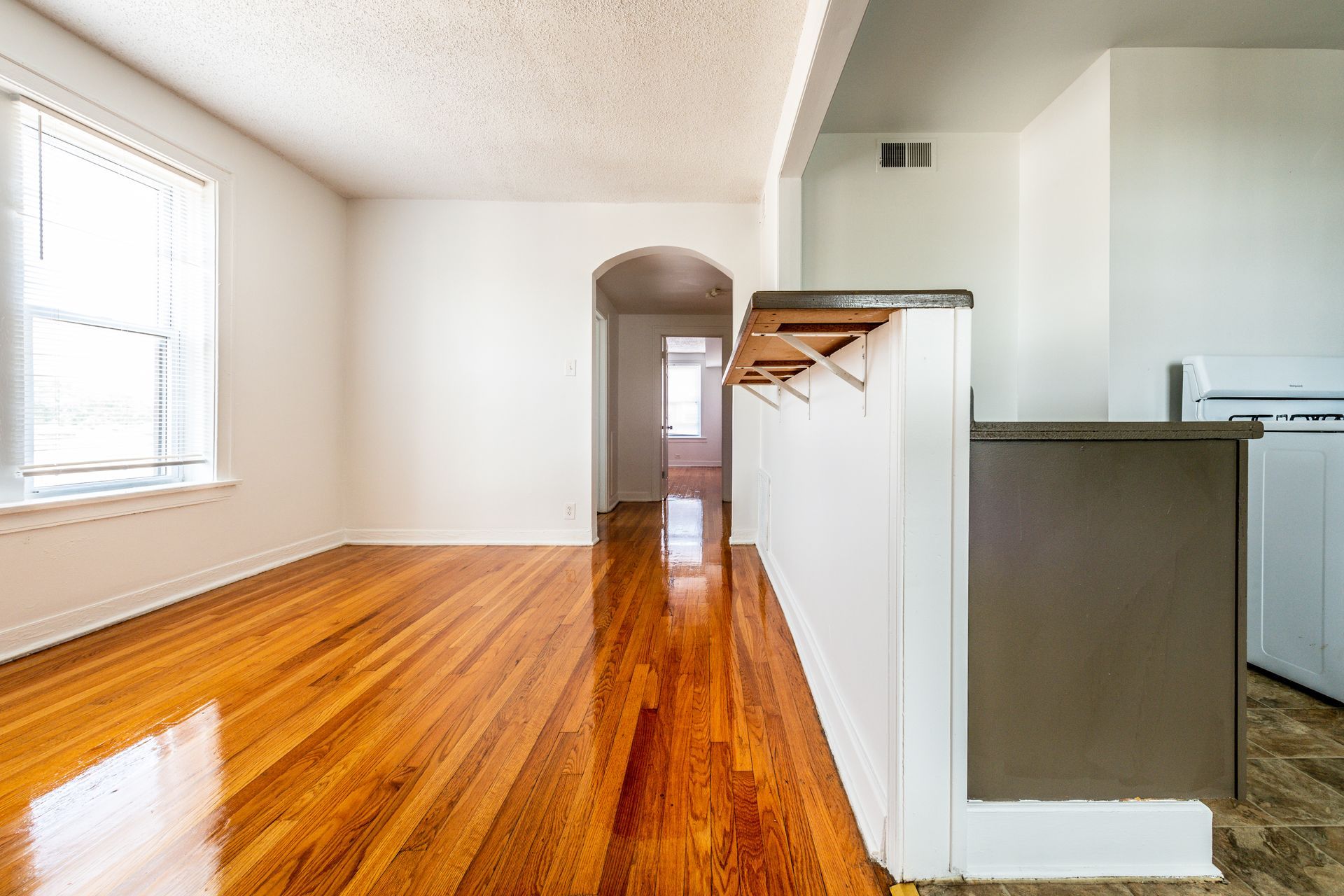 Empty apartment interior with wood floors, white walls, and a hallway.