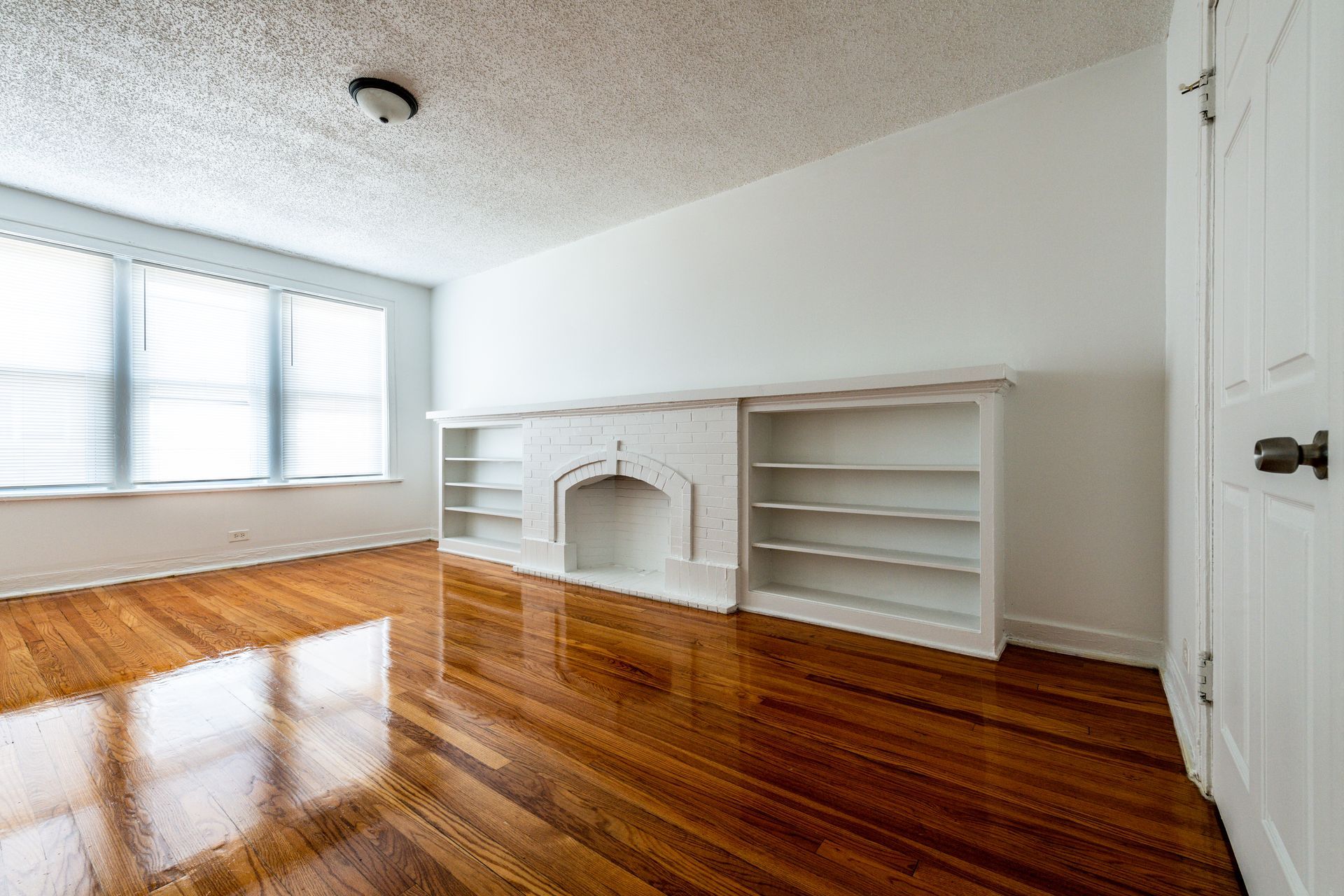 Empty room with hardwood floors, white walls, and built-in fireplace and bookshelves. Window on the left.