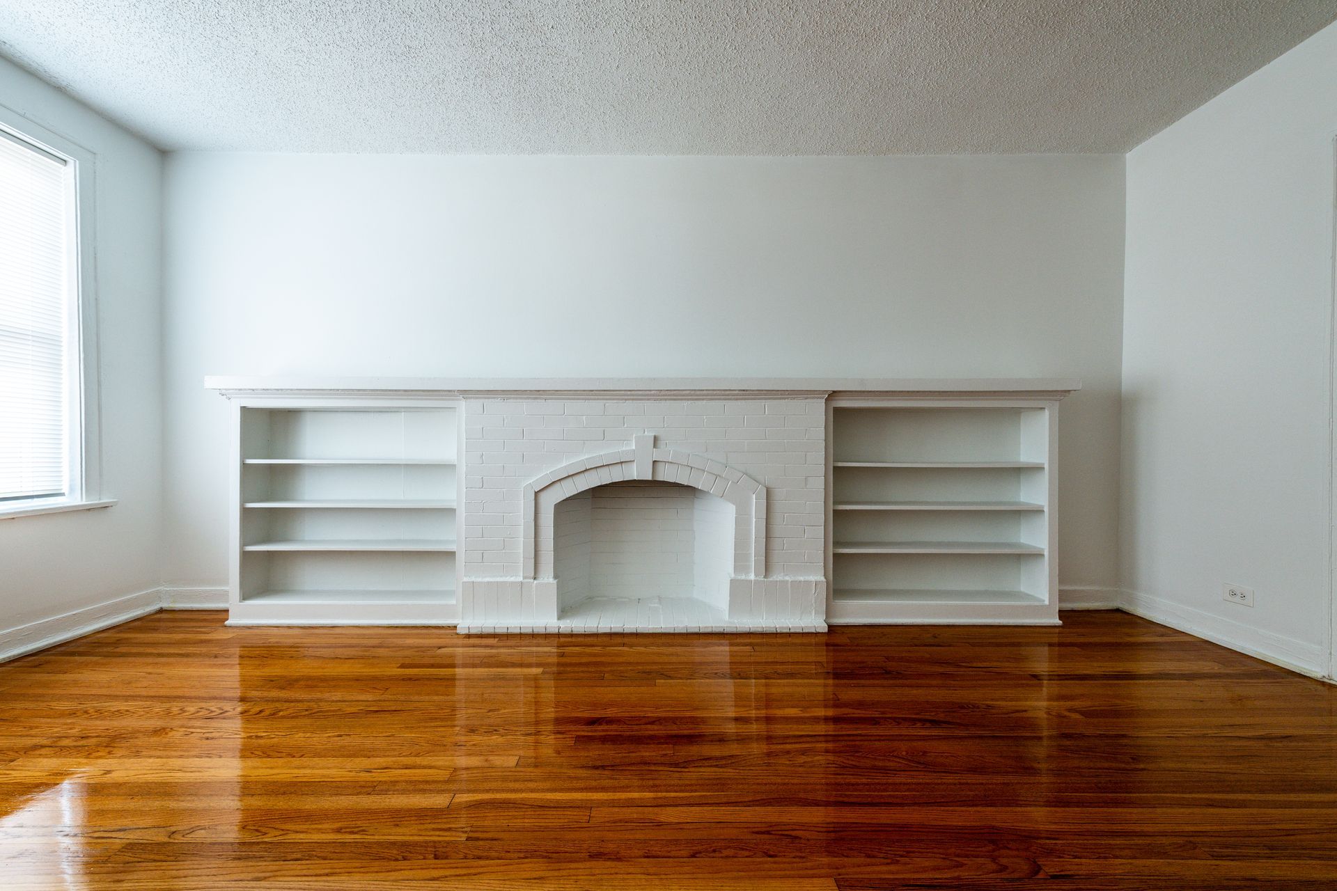 Empty living room with wood floors, white fireplace, built-in shelves, and a window.