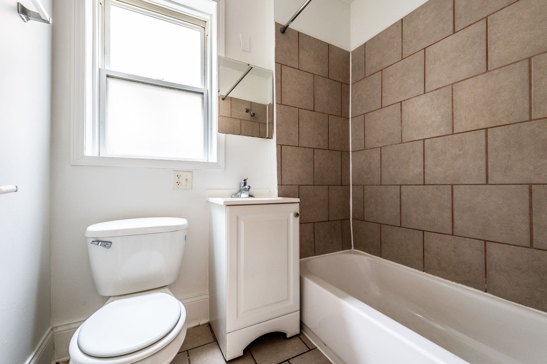 Bathroom with toilet, vanity, and bathtub with brown tile surround; window provides natural light.