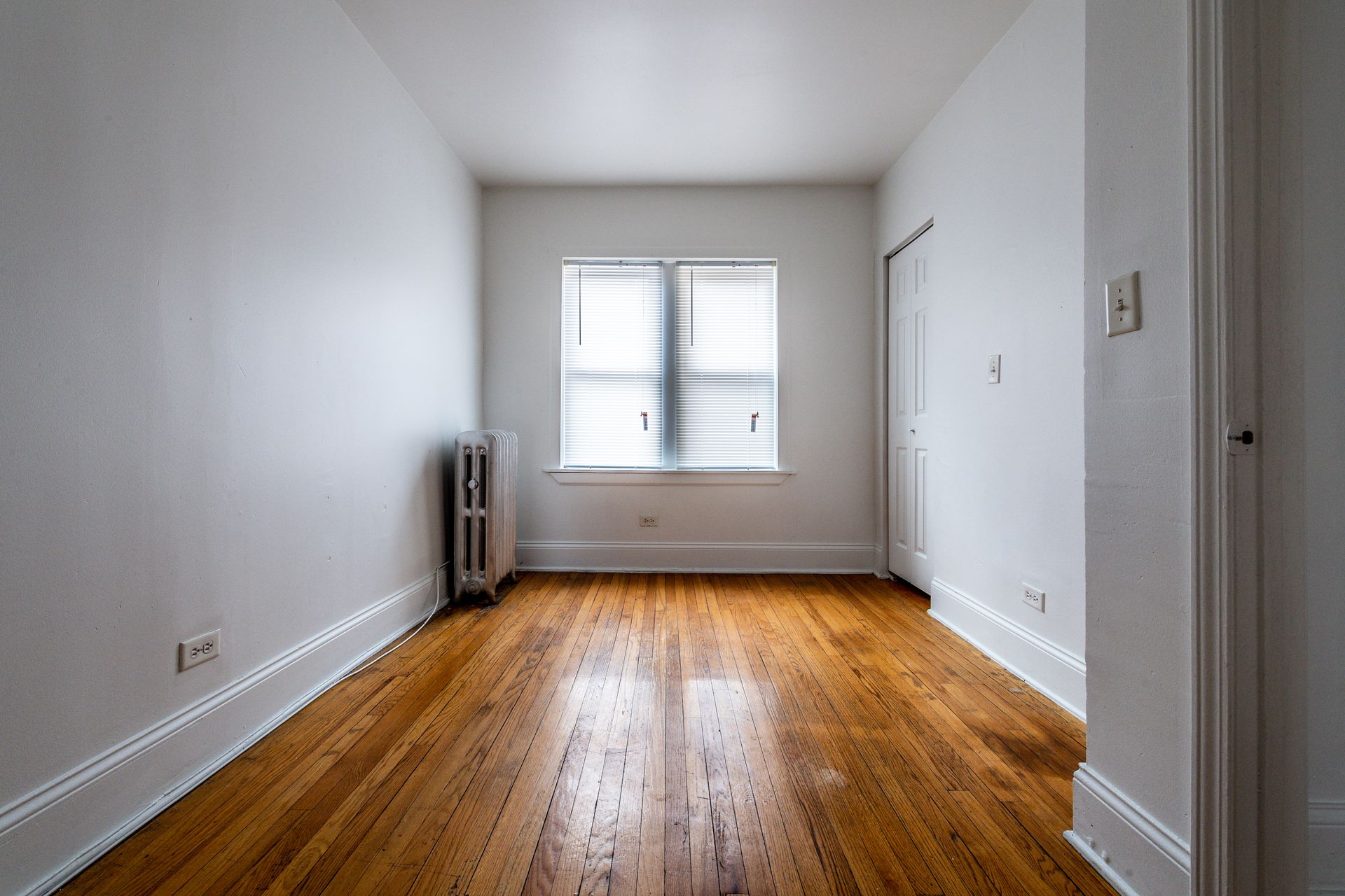 Empty room with hardwood floors, a window, radiator, and a closet. White walls and trim.