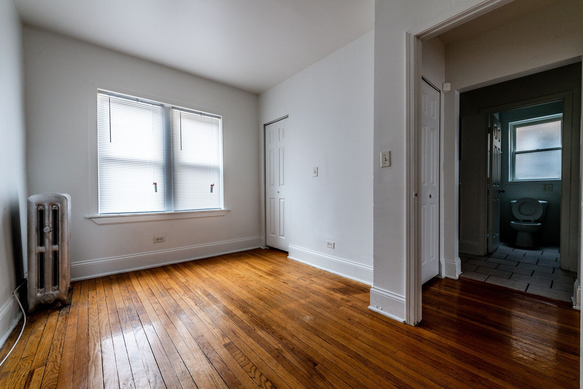 Empty room with hardwood floors, window with blinds, and doorway to bathroom.