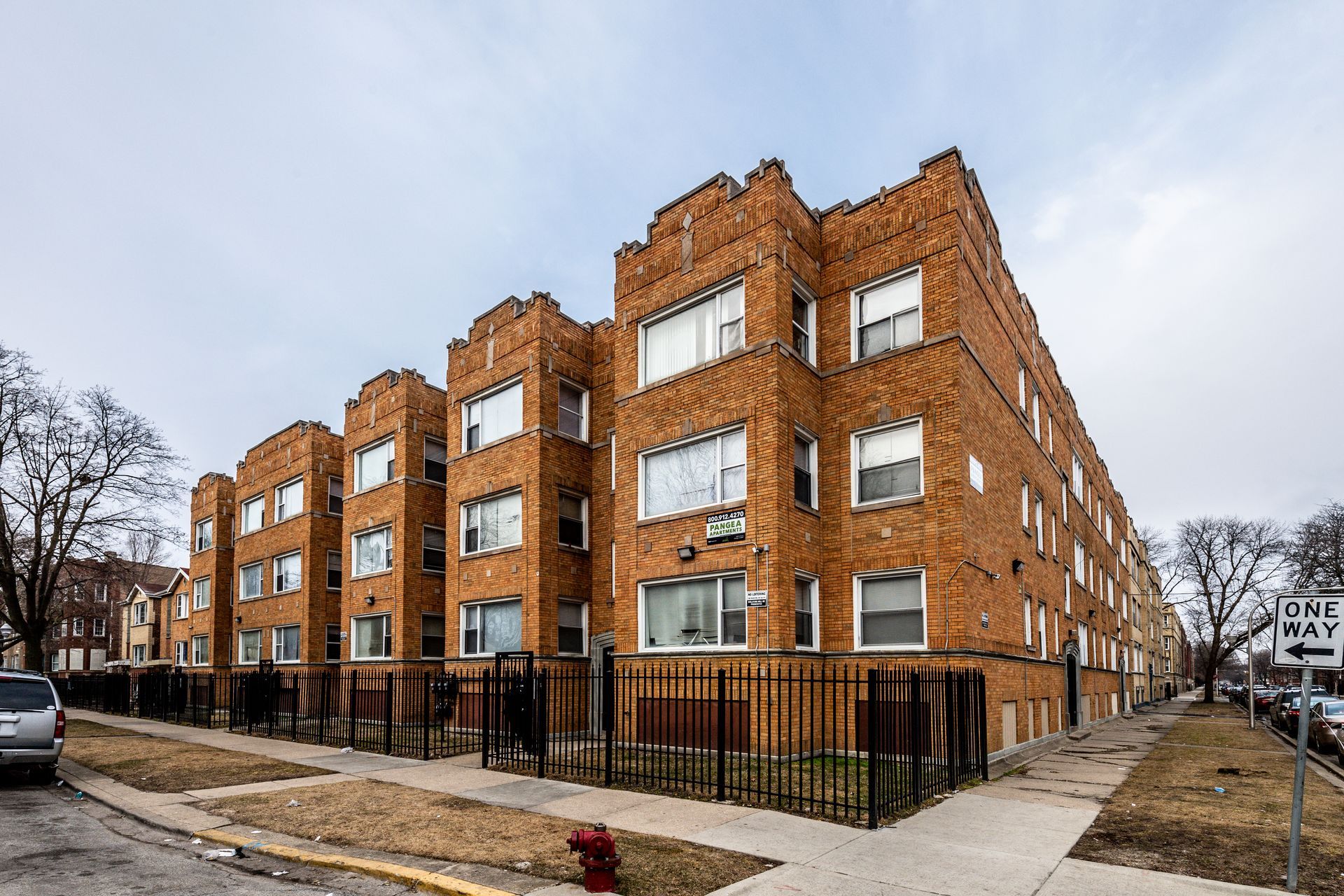 Multi-story brick apartment building on a residential street. Cloudy sky, brown fence.