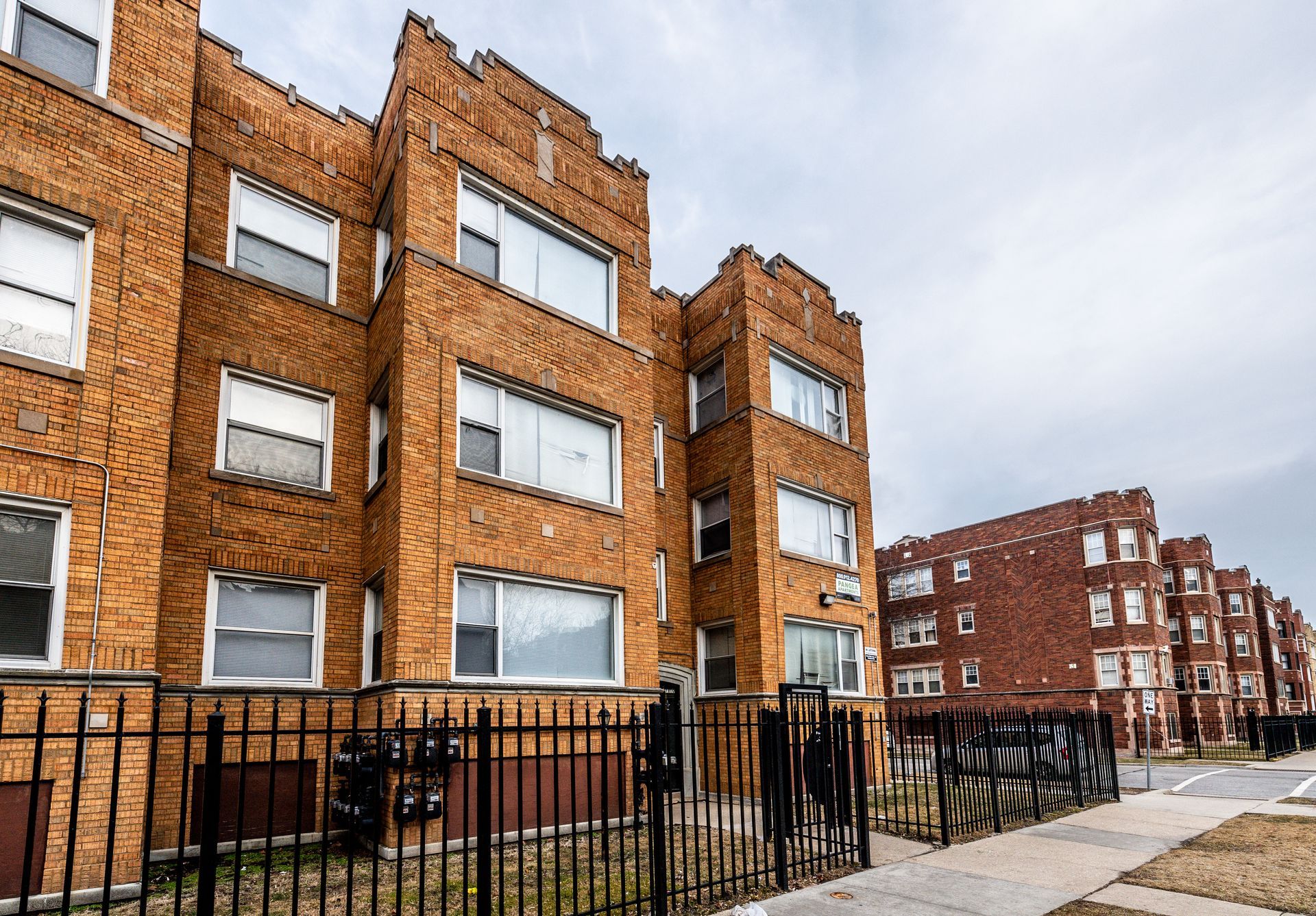 Multi-story brick apartment buildings with white-framed windows, behind a black metal fence, under a cloudy sky.