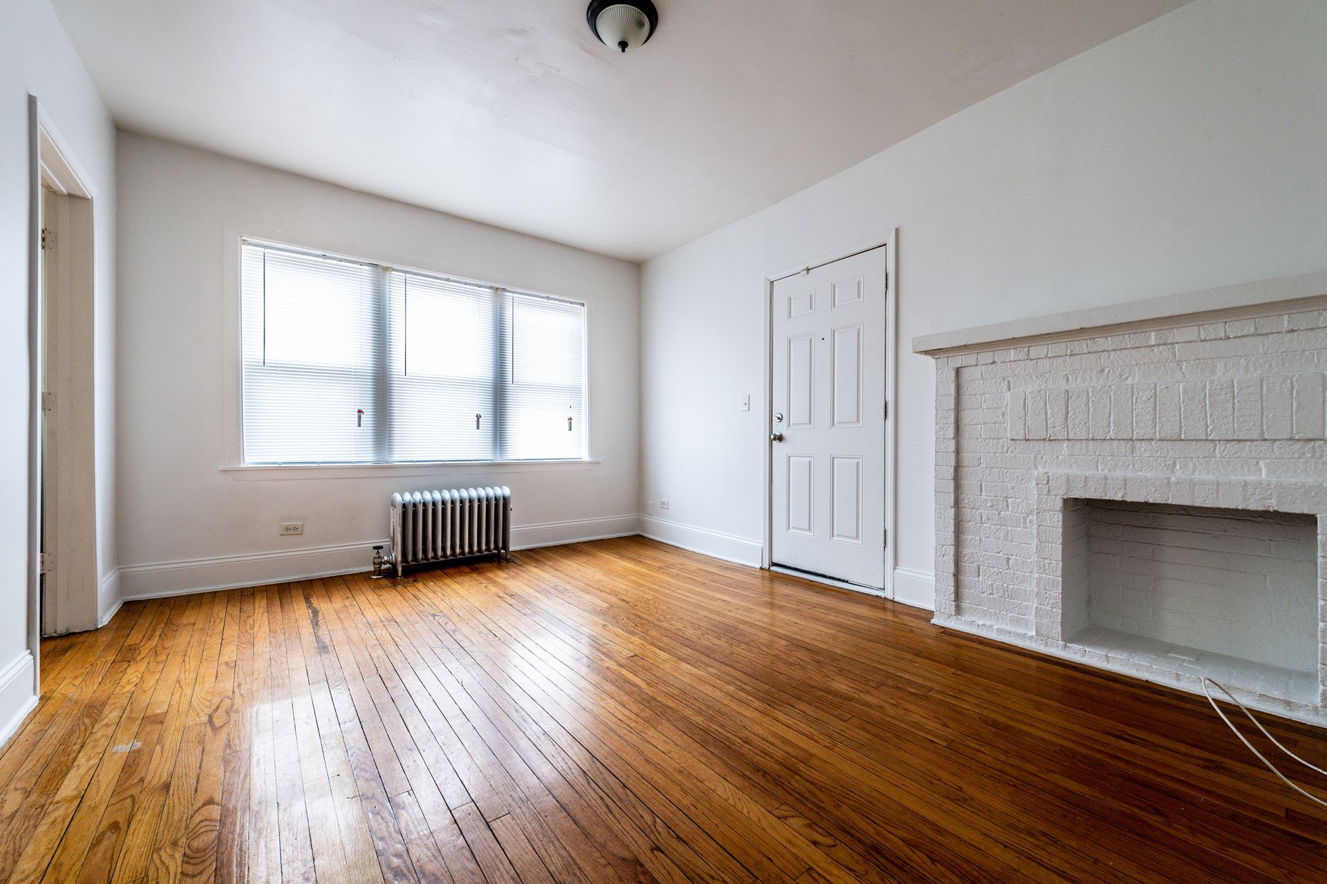 Empty room with hardwood floors, white walls, and a fireplace. Window with blinds and a radiator.