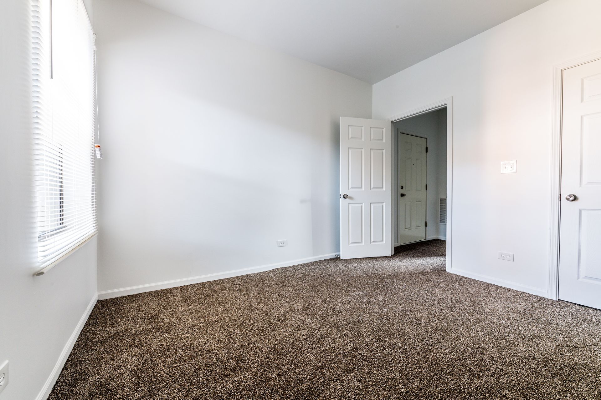 Empty room with brown carpet, white walls, and two white doors. A window with blinds is on the left.
