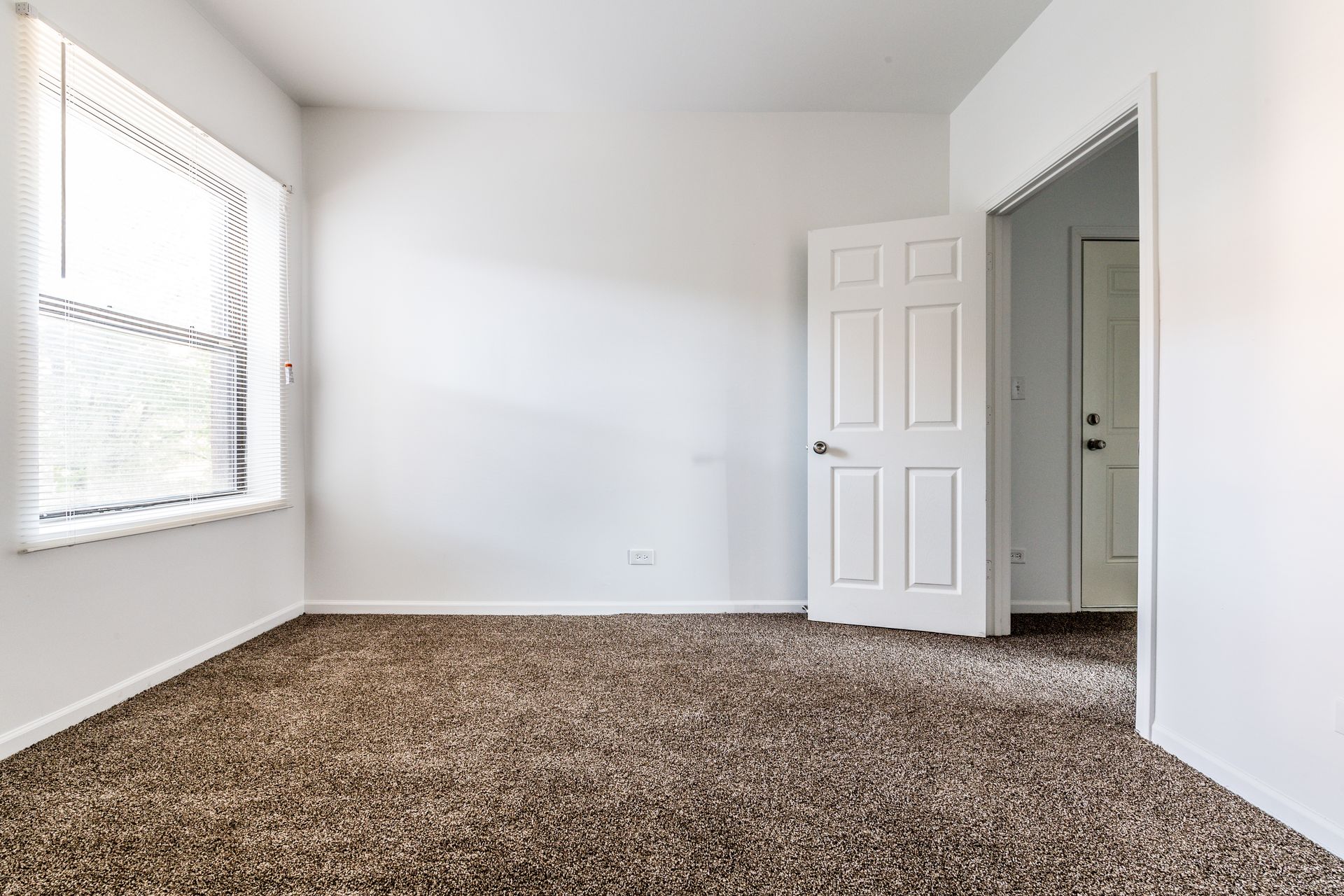 Empty room with white walls, brown carpet, window with blinds, and open white door.