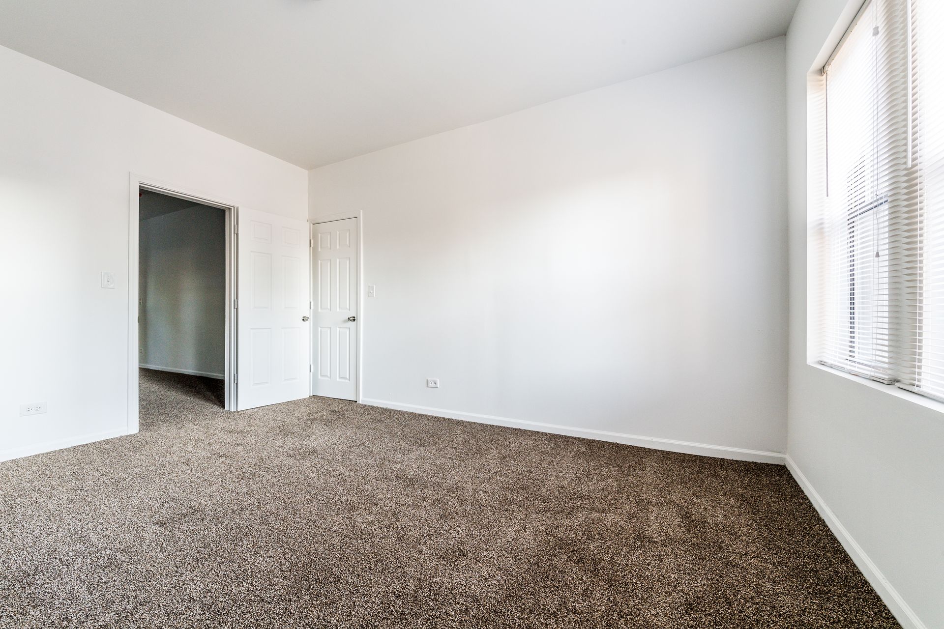 Empty room with brown carpet, white walls, two doors, and a window with blinds.