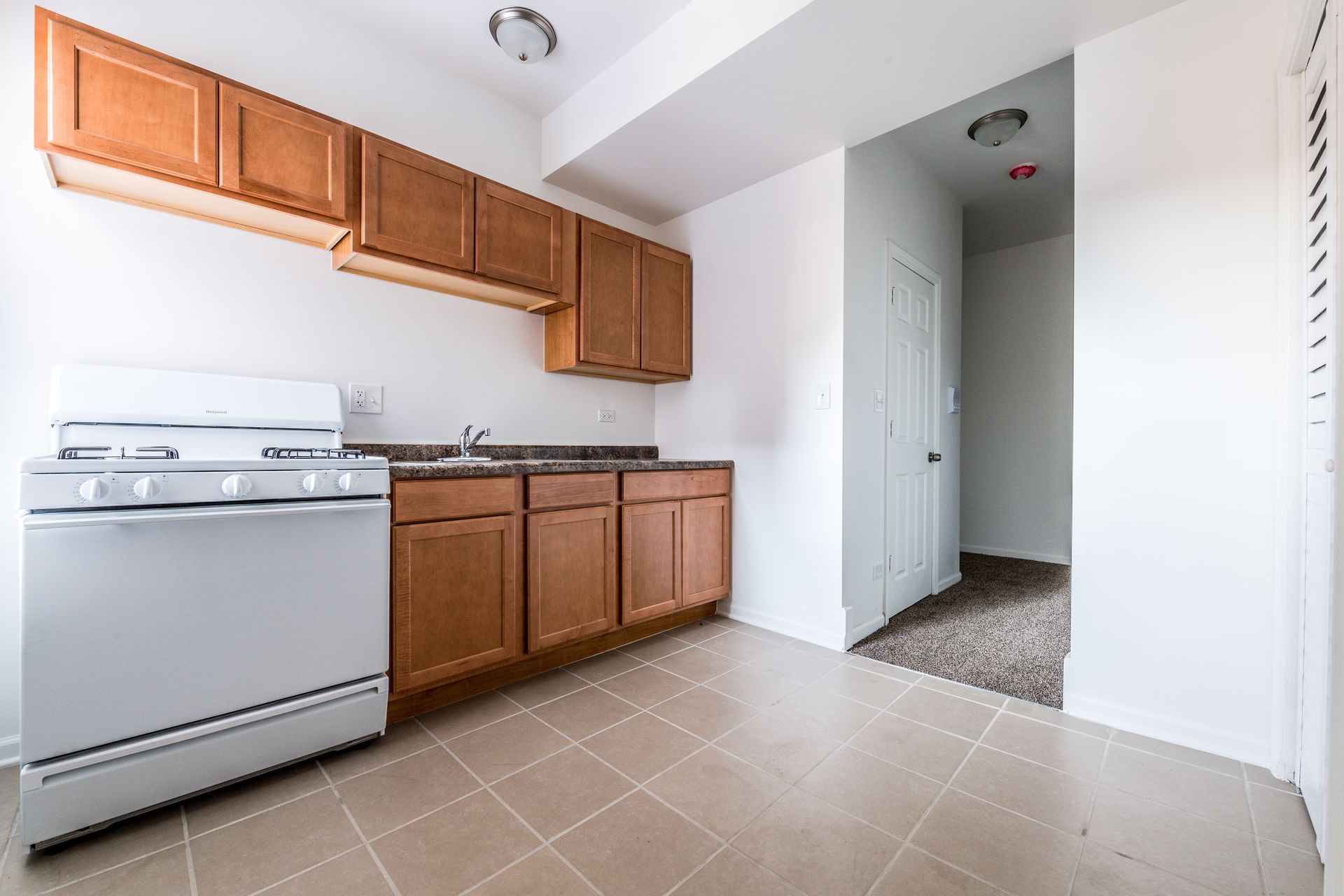 Small kitchen with wooden cabinets, white stove, and beige tile floor. Doorway leads to hallway.