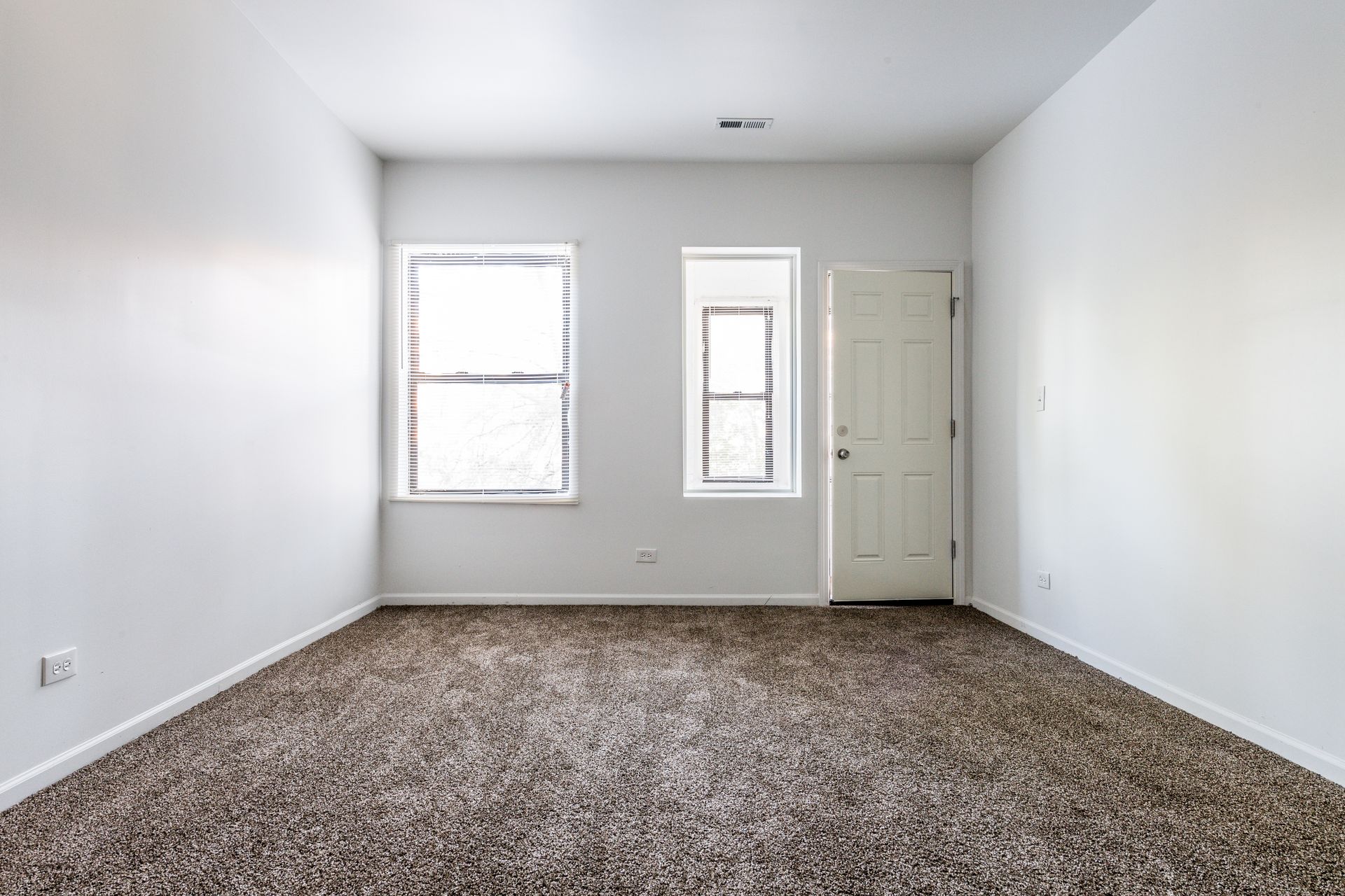 Empty room with brown carpet, white walls, two windows, and a door.