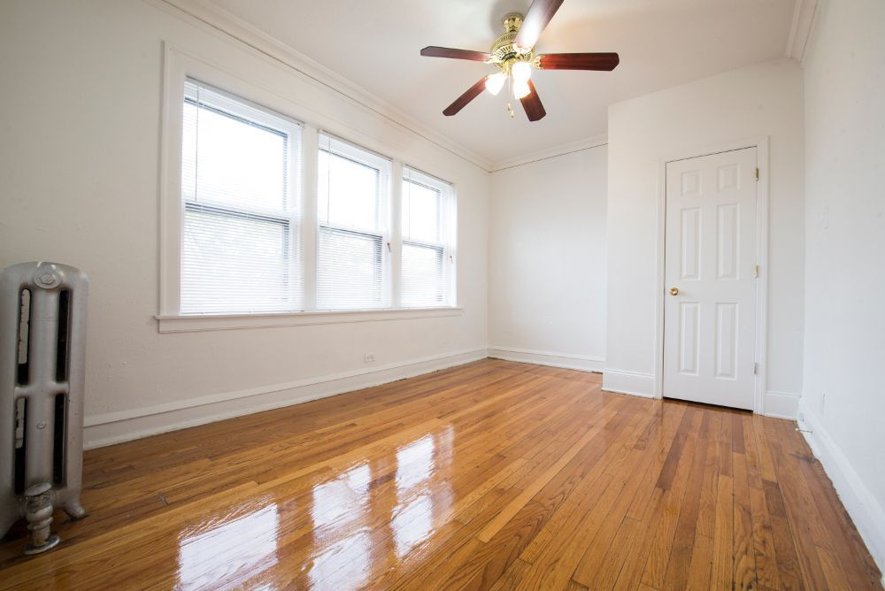 Empty room with hardwood floors, a radiator, and a ceiling fan; windows and a closed door are also present.