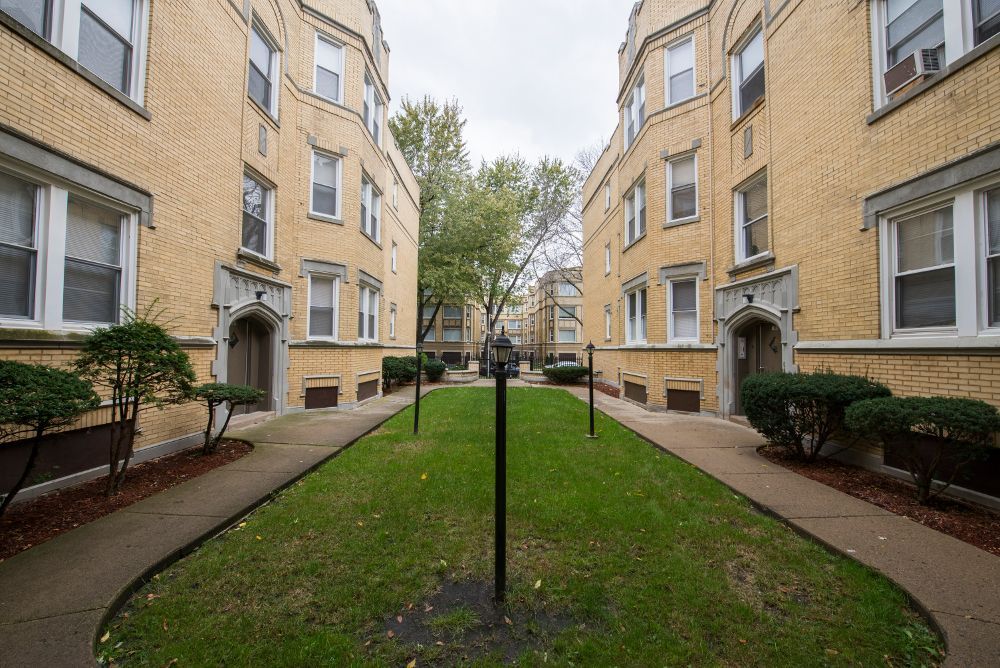 Two yellow brick apartment buildings flank a green grassy courtyard with a central path.