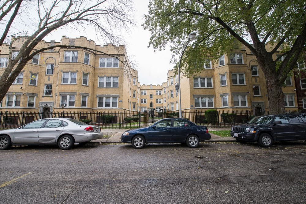 Apartment buildings with cars parked on a street in front of them, trees frame the view, cloudy sky.