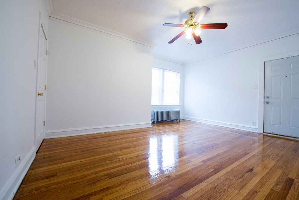 Empty room with wooden floors, white walls, ceiling fan, and a closed door.