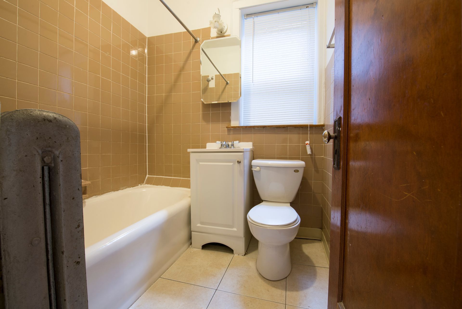 Small bathroom with tan tiled walls, white tub, toilet, and vanity, and a closed brown door.