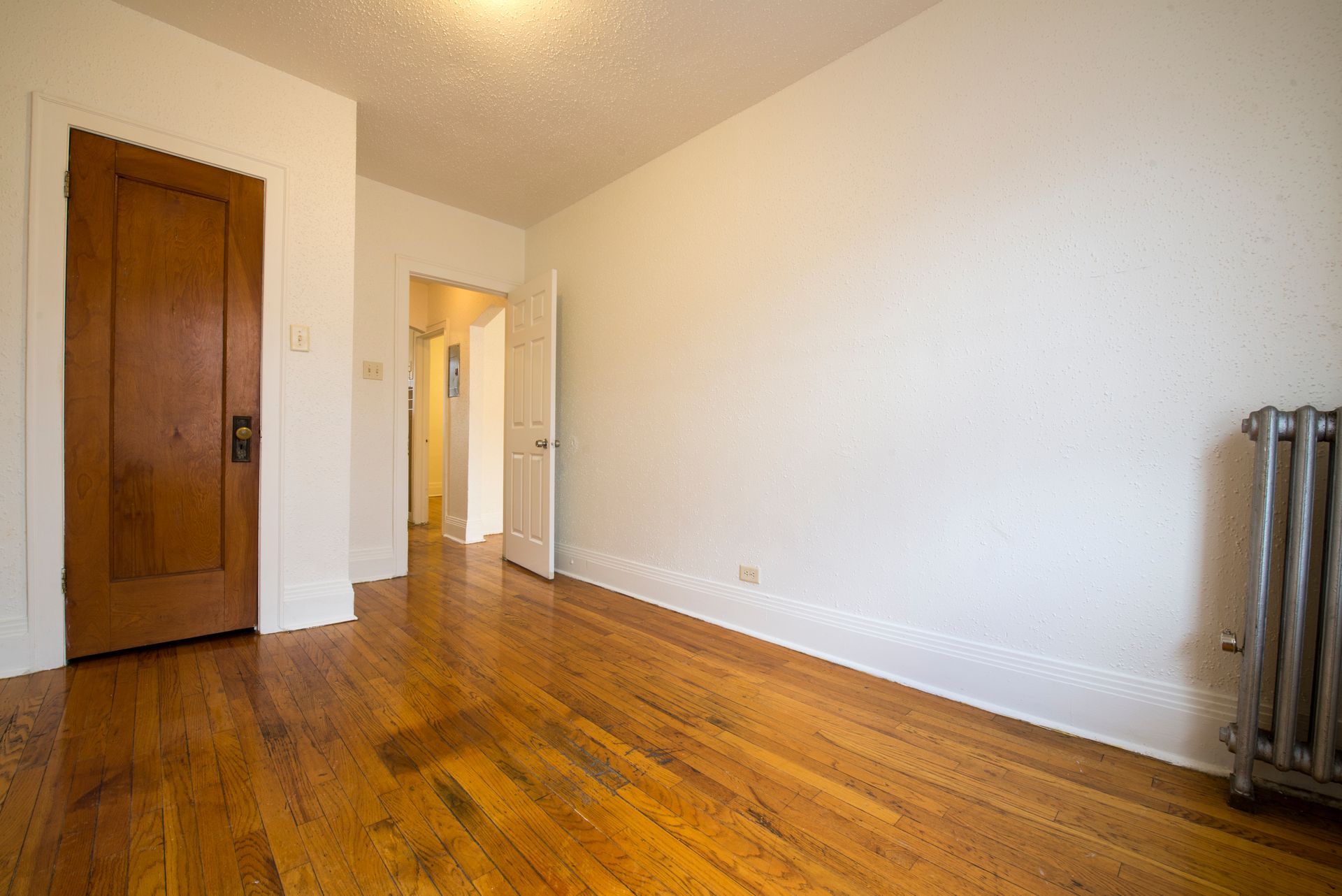 Empty room with hardwood floors, white walls, and a radiator. A wooden door is on the left.