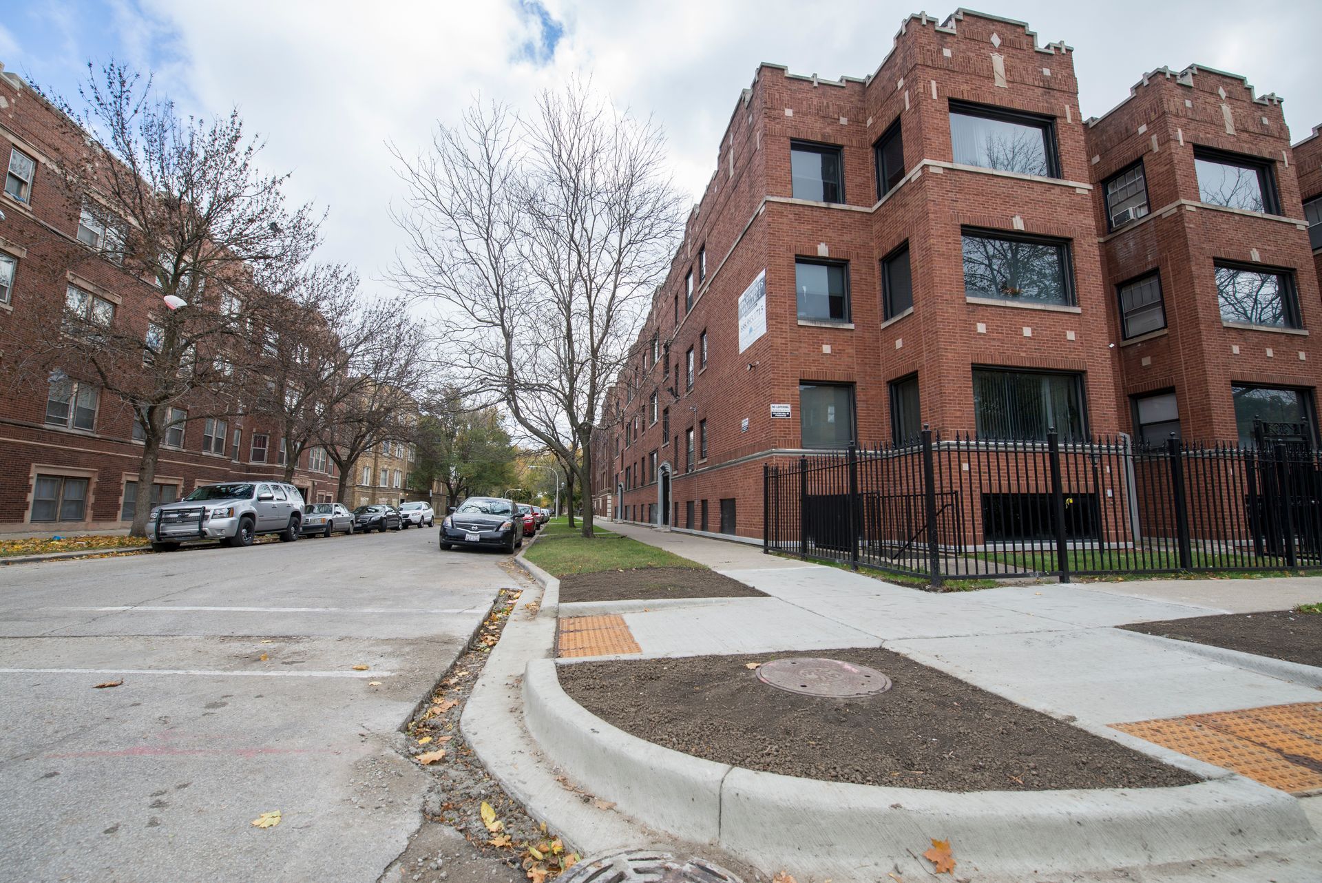 Brick apartment buildings line a street. Cars are parked along the side, curb and sidewalk in foreground.