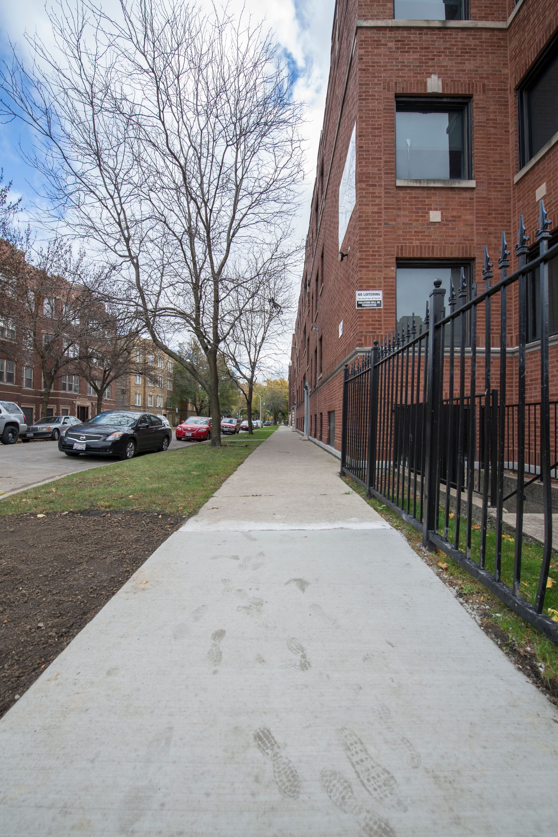 Sidewalk next to brick building with black fence and a tree, parked cars on the left.