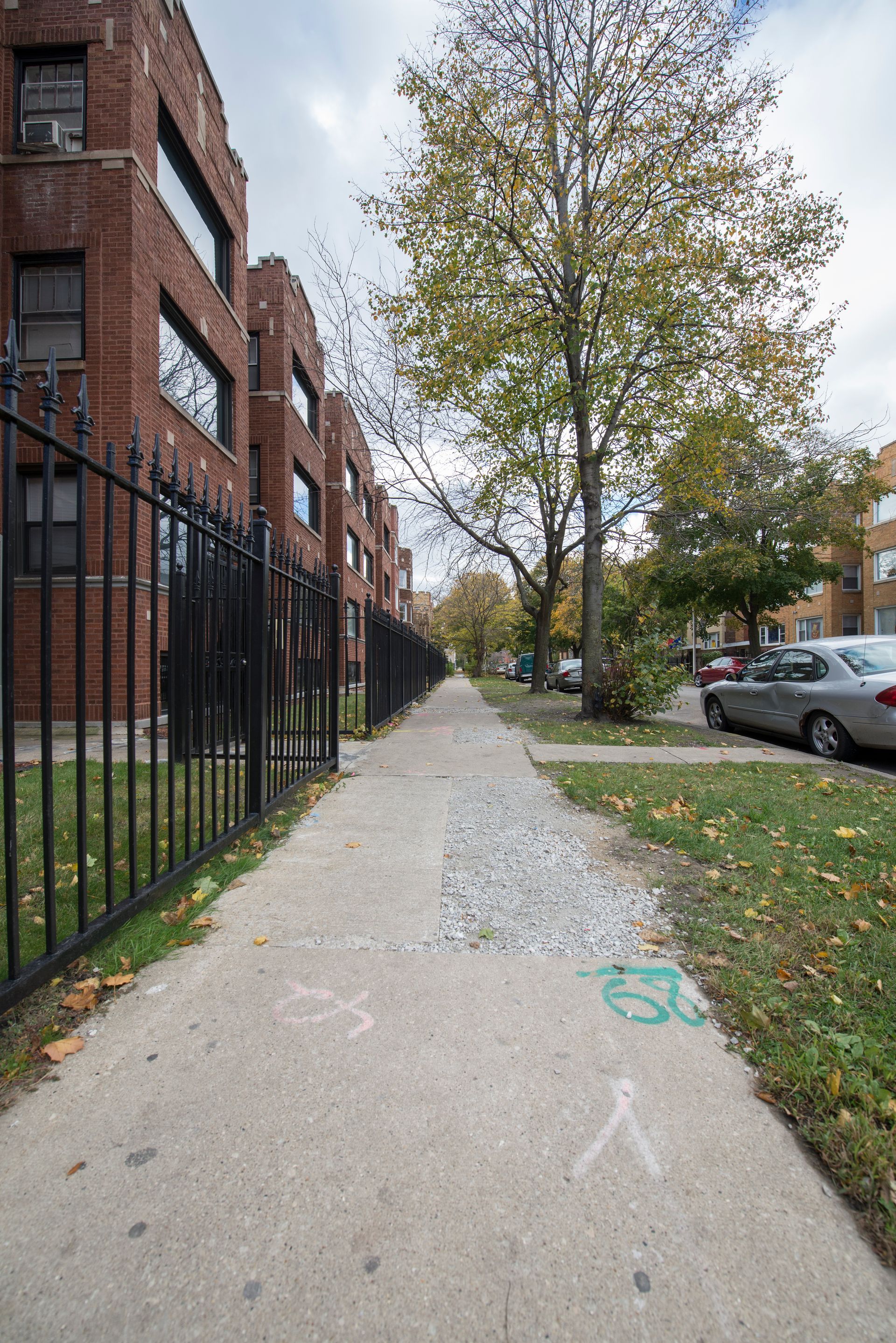 Sidewalk alongside brick buildings and a black metal fence, leading to trees and a street with parked cars.