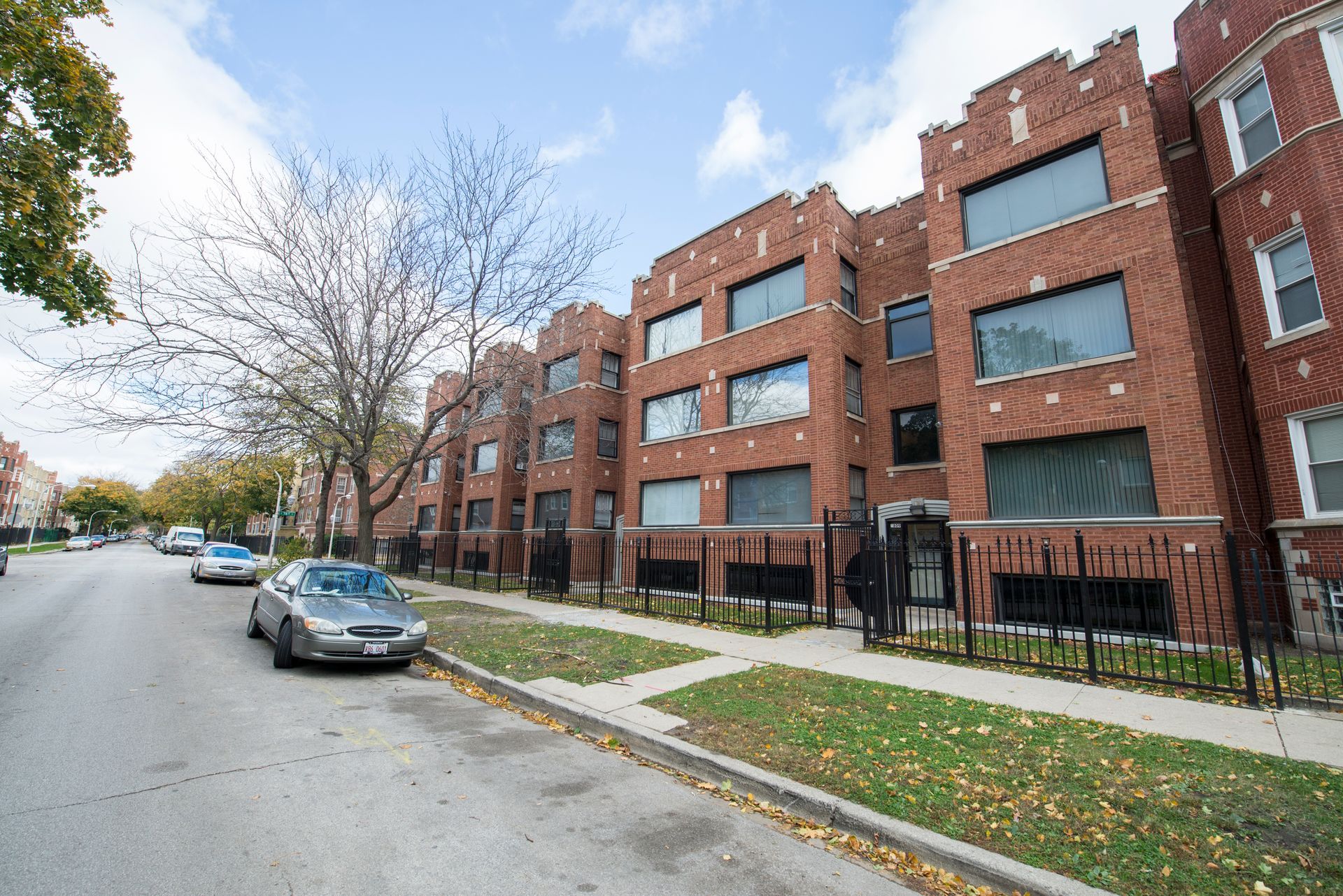 Brick apartment building on a city street, with parked cars and a tree with bare branches.