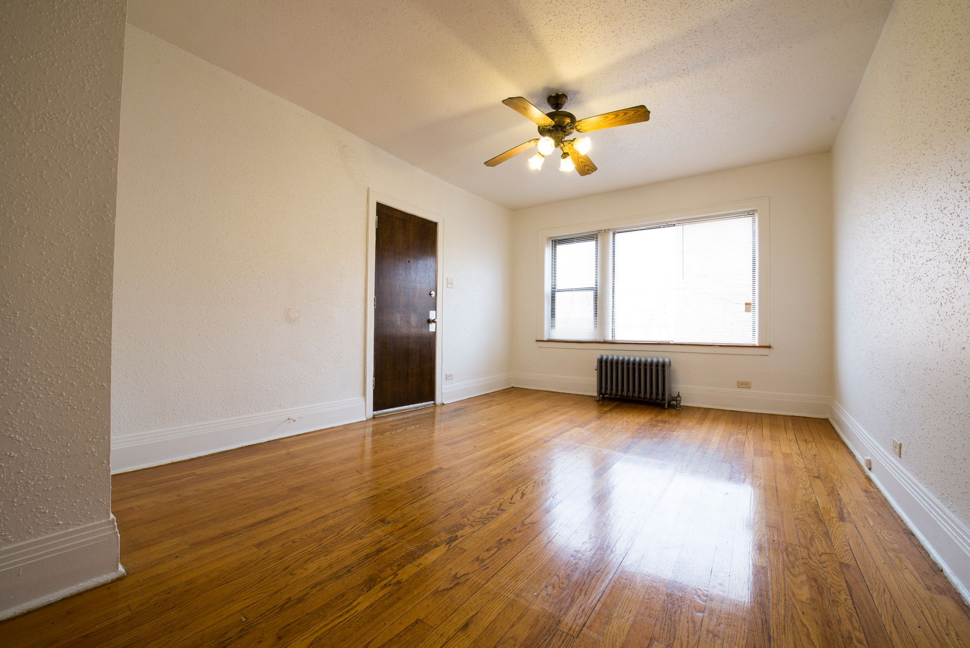 Empty room with hardwood floors, a closed brown door, and a window with a radiator. Ceiling fan present.