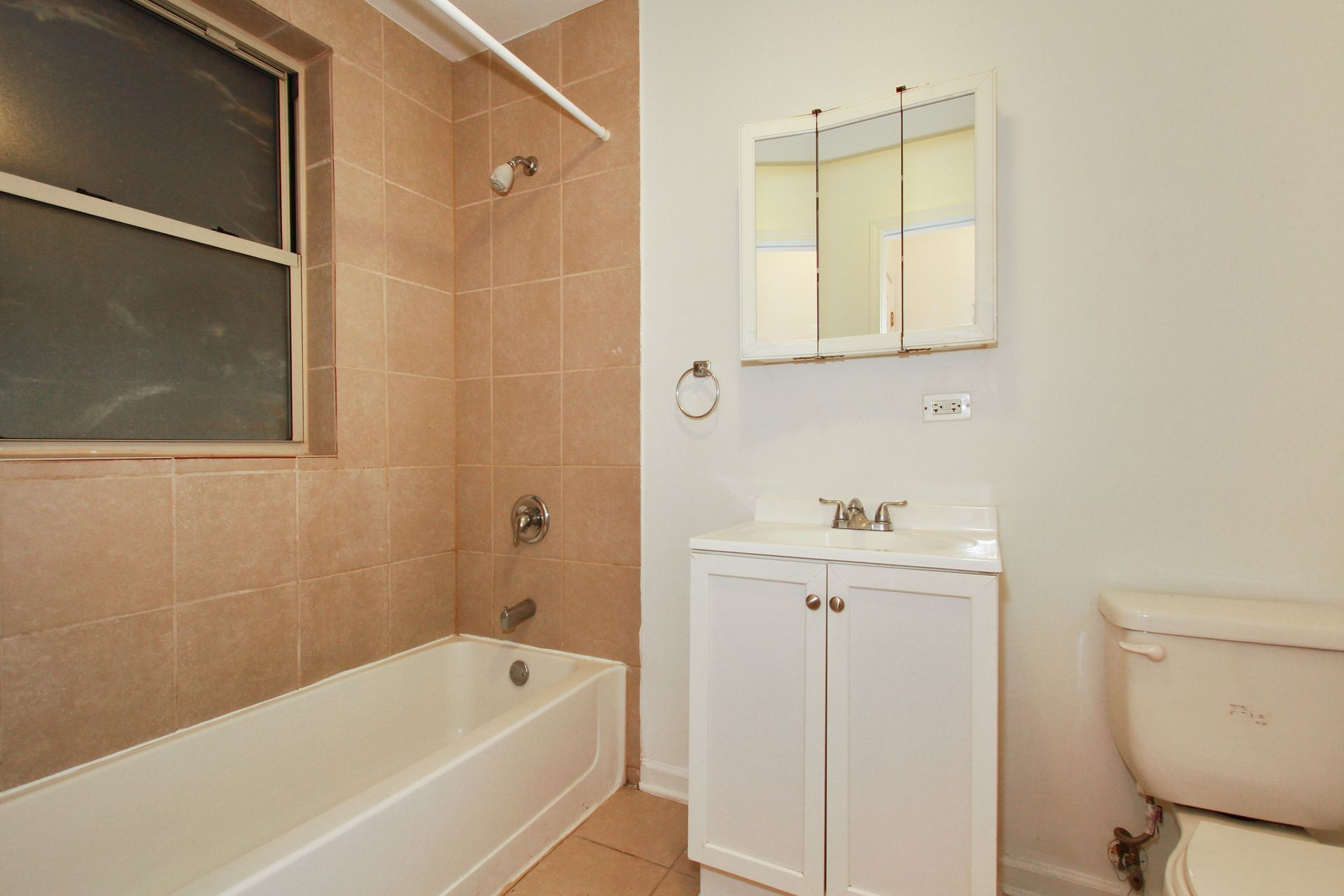 Bathroom with a white bathtub, toilet, and vanity. Beige tile, window, and mirrored cabinet.
