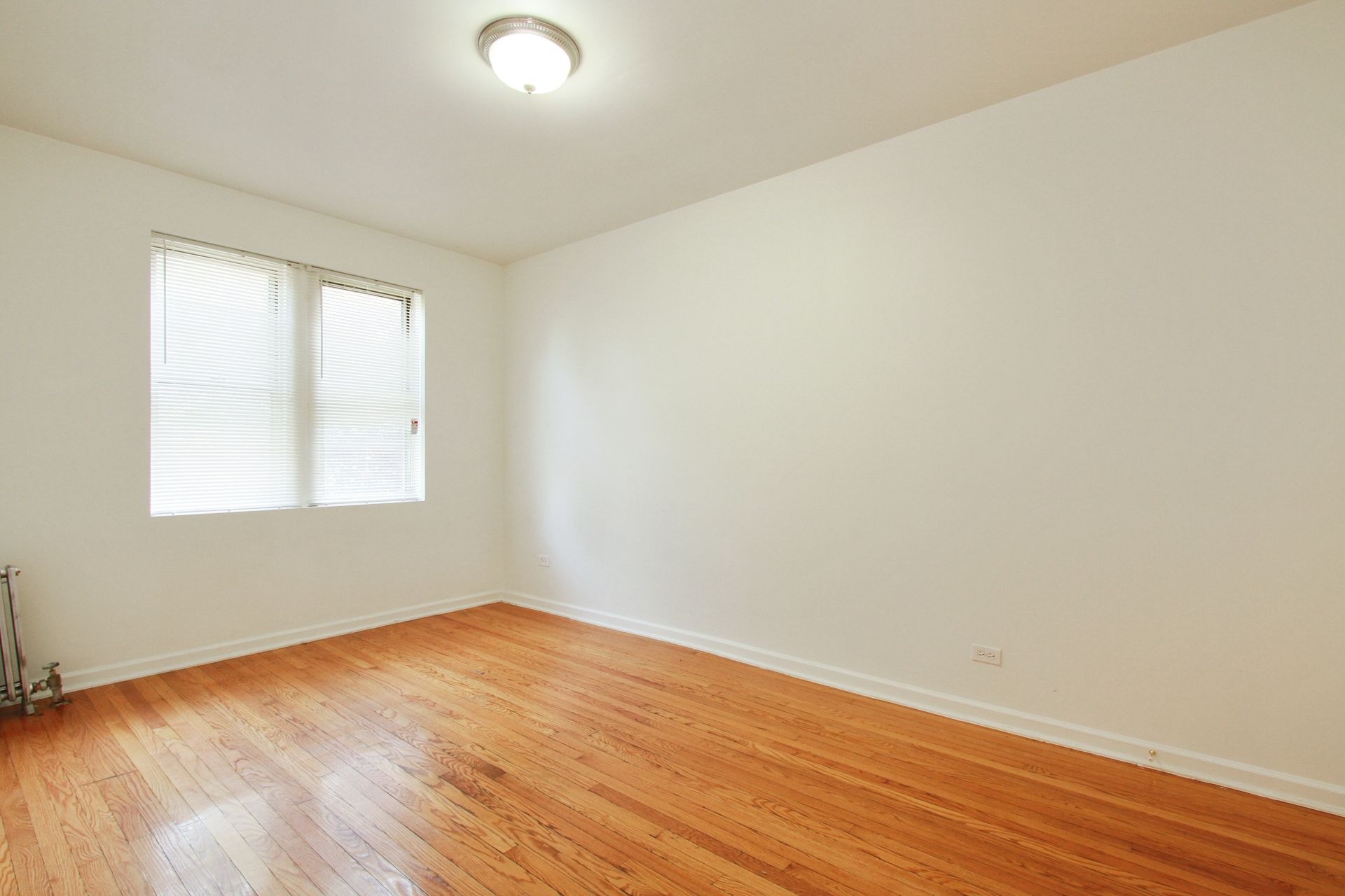 Empty room with hardwood floors, a window with blinds, and a ceiling light.
