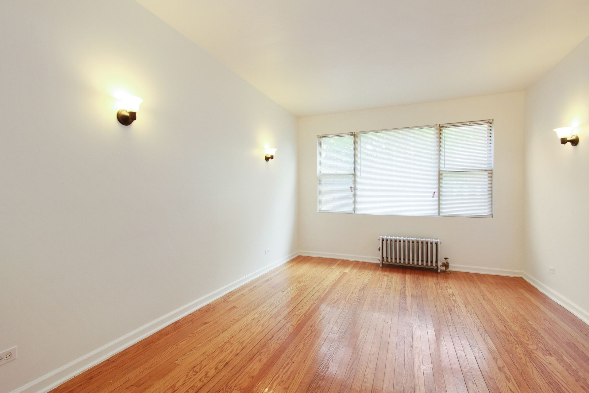 Empty room with hardwood floors, white walls, three sconces, and a window with blinds.
