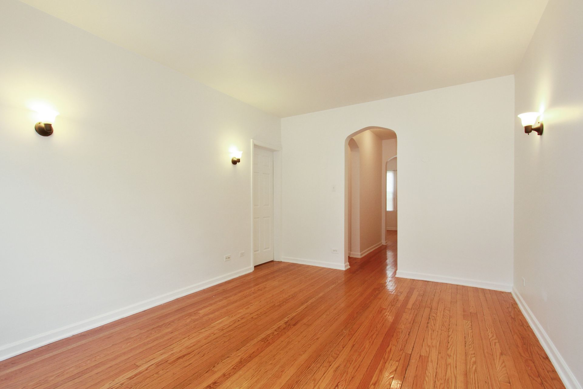 Empty room with hardwood floors, white walls, arched doorway, sconce lighting.